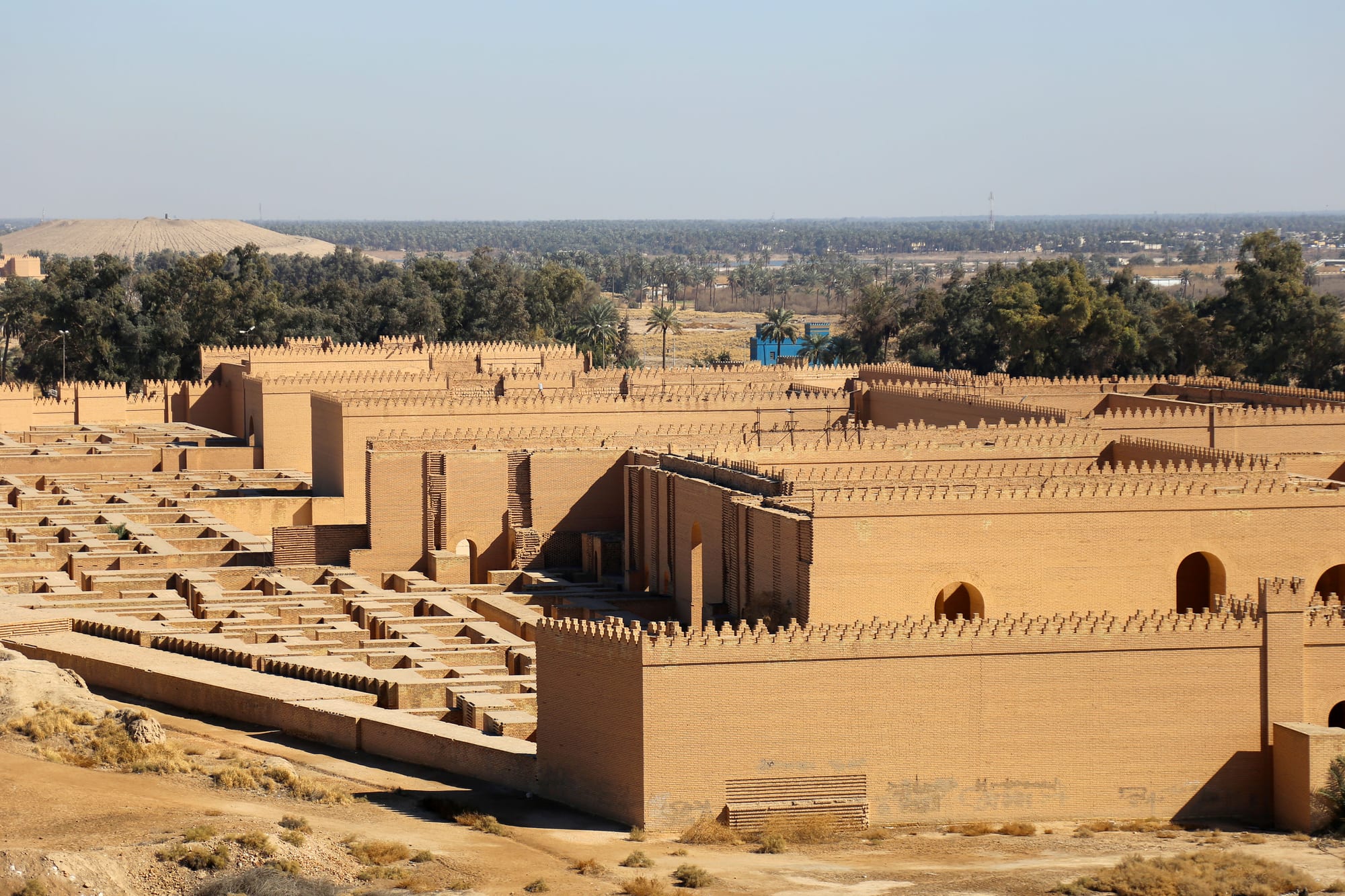 Long walls of pale brown brick with crenellated tops and arched doorways, rebuilt over the ruins of an ancient city, with palm groves behind