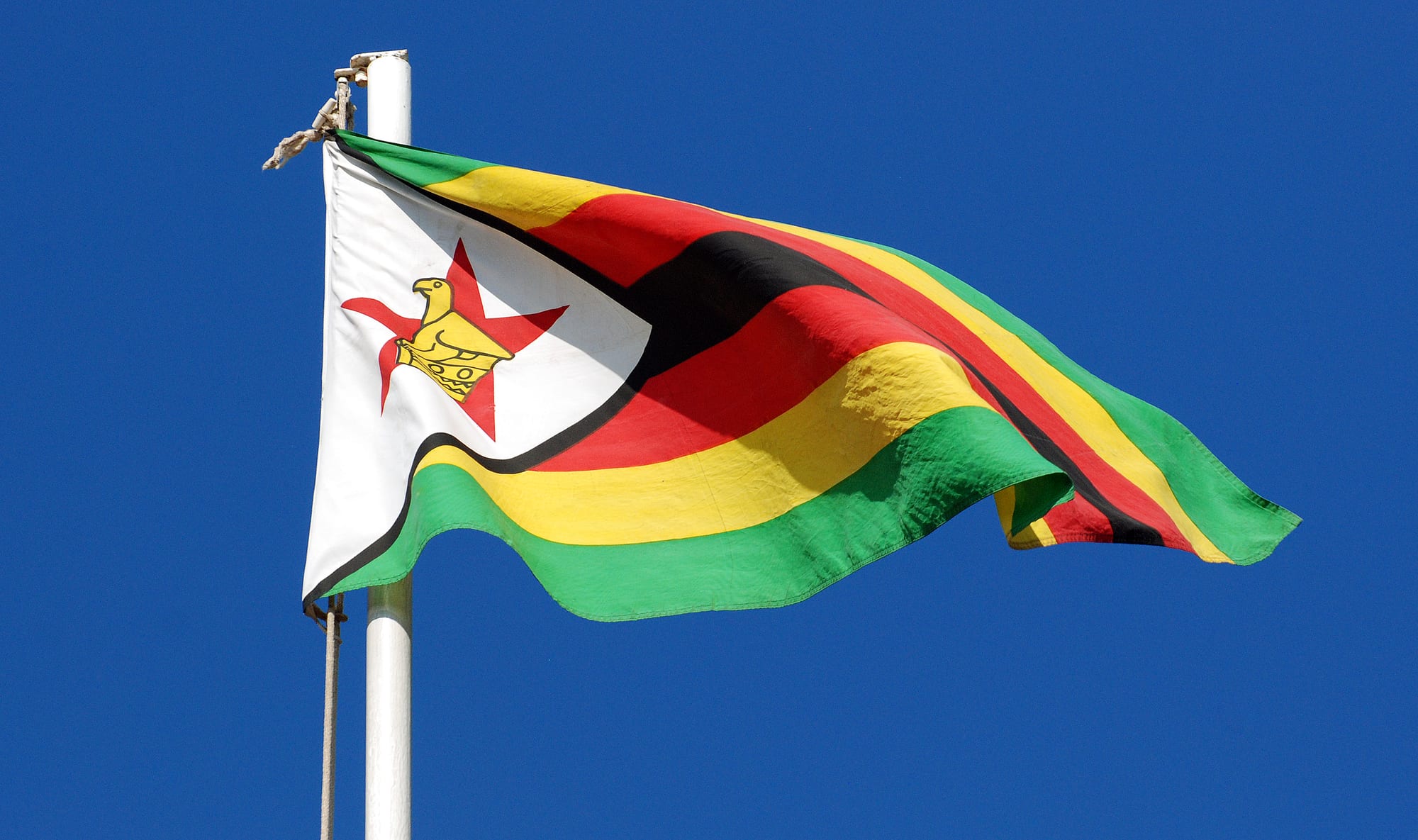 The flag of Zimbabwe, with green, yellow, red and black stripes and a gold bird on a red star in a white triangle, flying against a deep blue sky