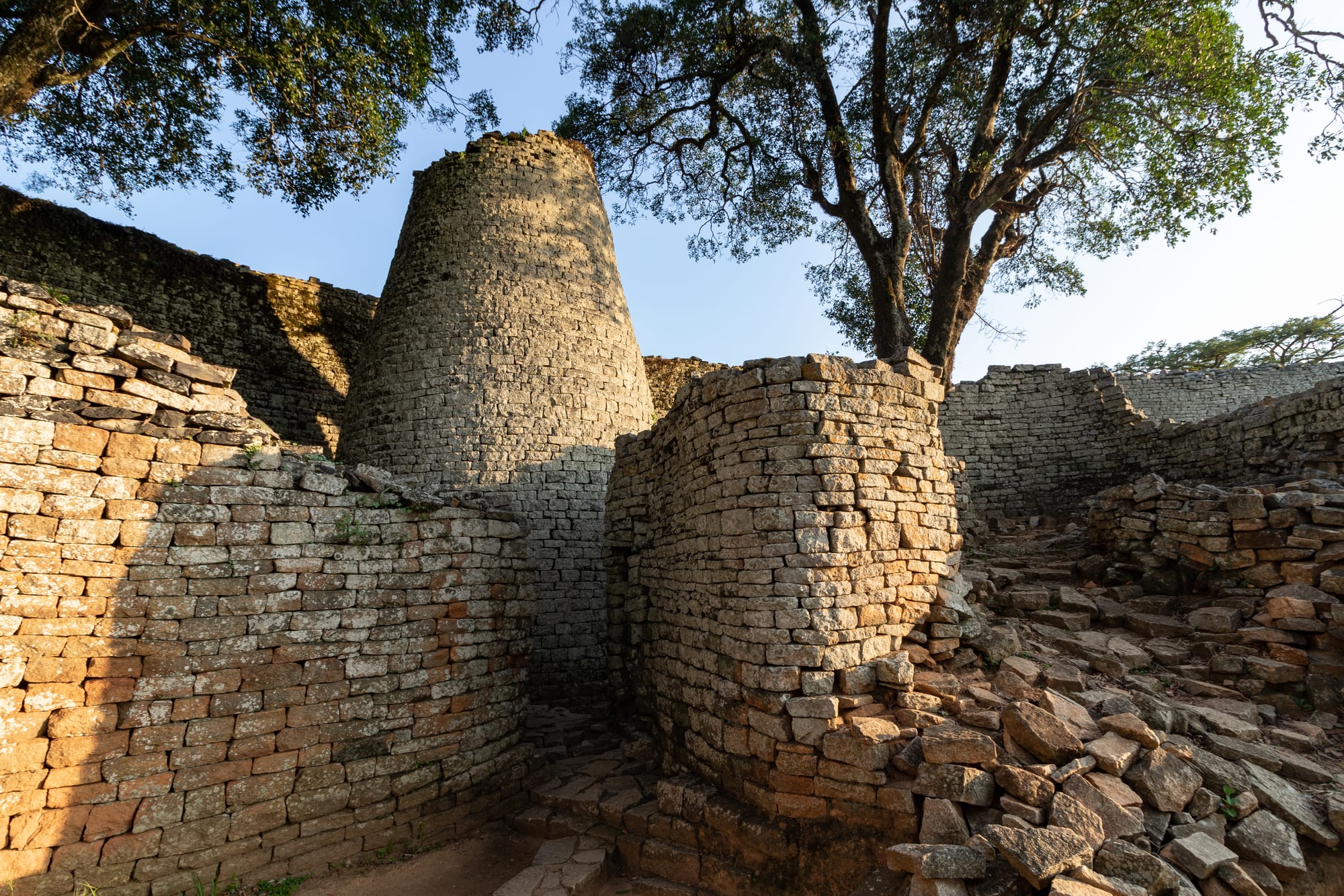 A tall, tapering tower of neatly stacked granite blocks between curving dry-stone walls, under trees and a blue sky