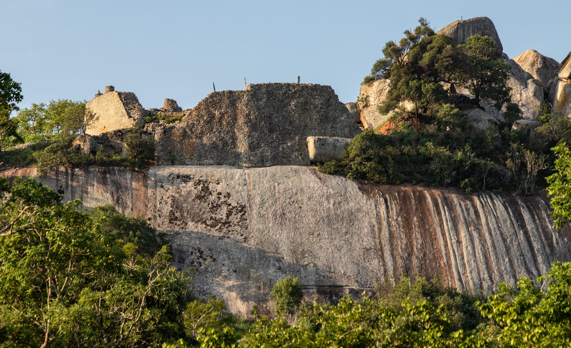 Ruined dry-stone walls built along the top of a huge, smooth granite cliff, among boulders and trees
