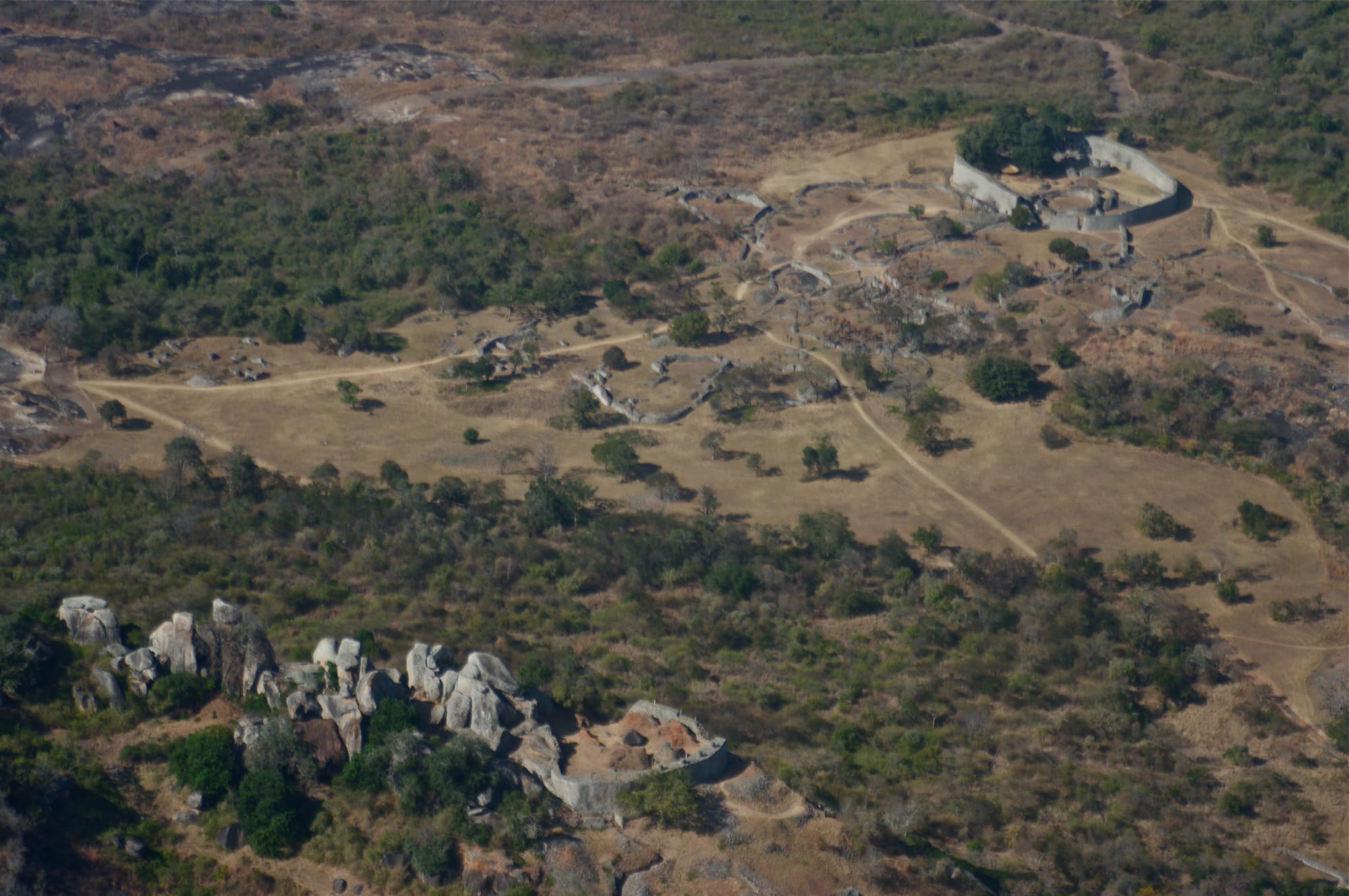 An aerial view over dry bush to an oval stone enclosure in a grassy valley, with ruined walls on a boulder-strewn hilltop in the foreground