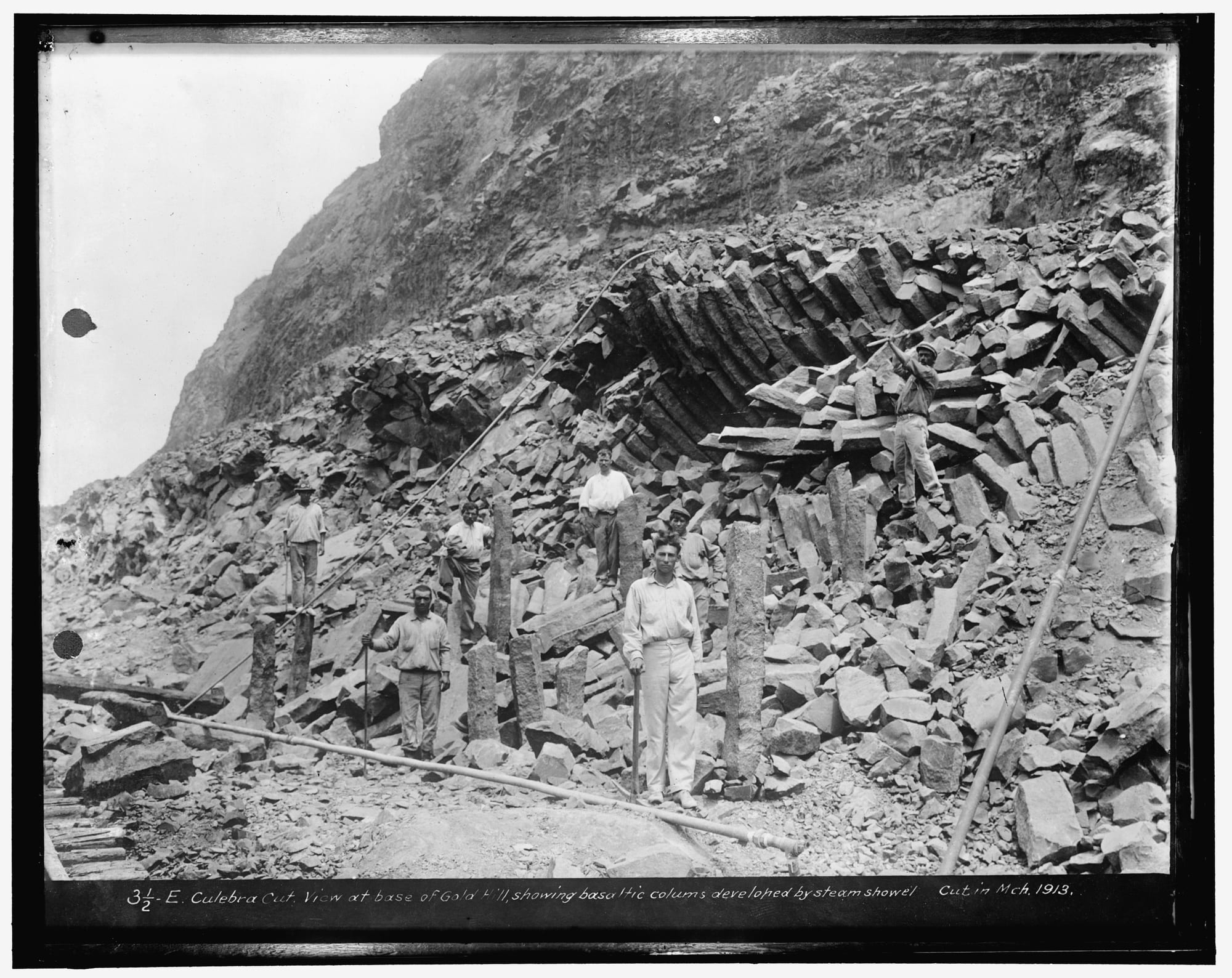 Workers with tools standing among broken columns of dark basalt rock at the foot of a steep rocky hill