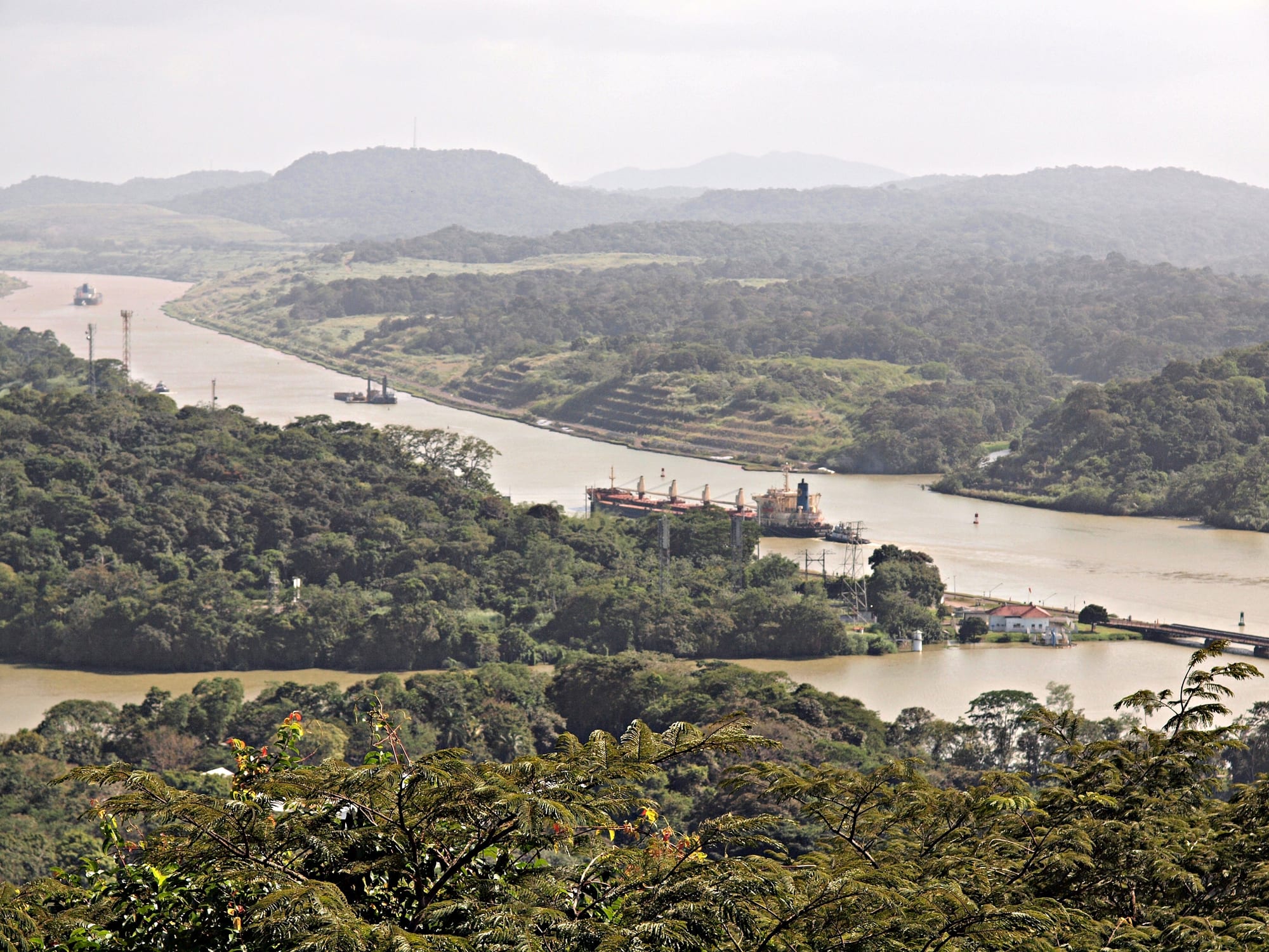A wide brown waterway winding between forested hills, with a cargo ship passing a small bridge