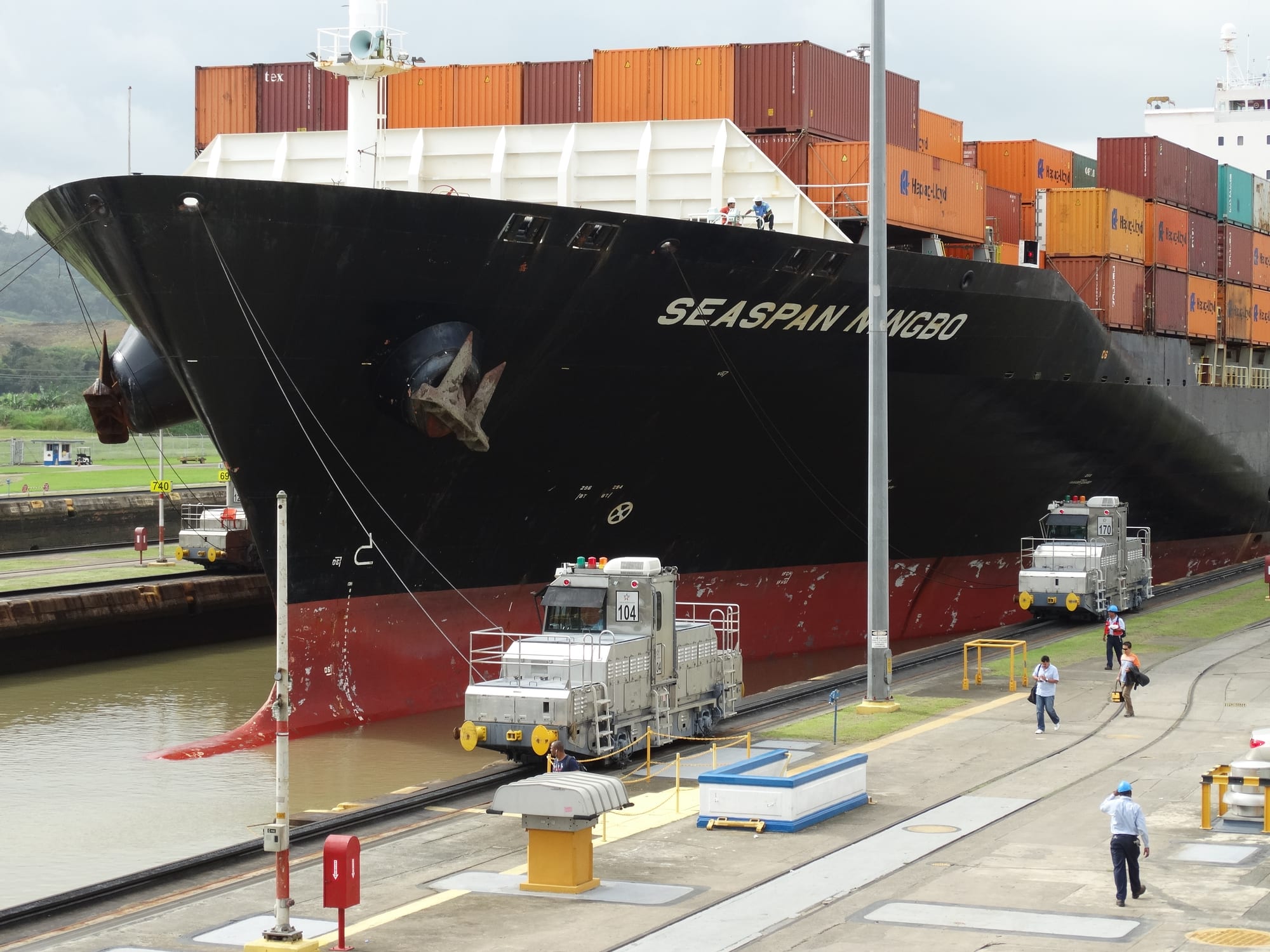 A large black container ship stacked with containers in a lock chamber, held on cables by small silver rail locomotives on the lock wall