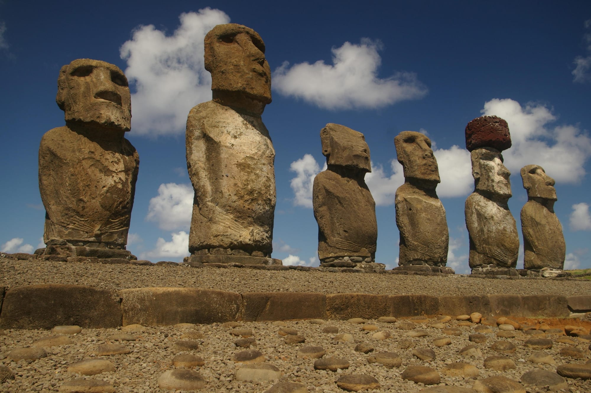Six weathered stone statues with long faces and full torsos standing in a row on a stone platform under a blue sky, one wearing a red stone cylinder on its head