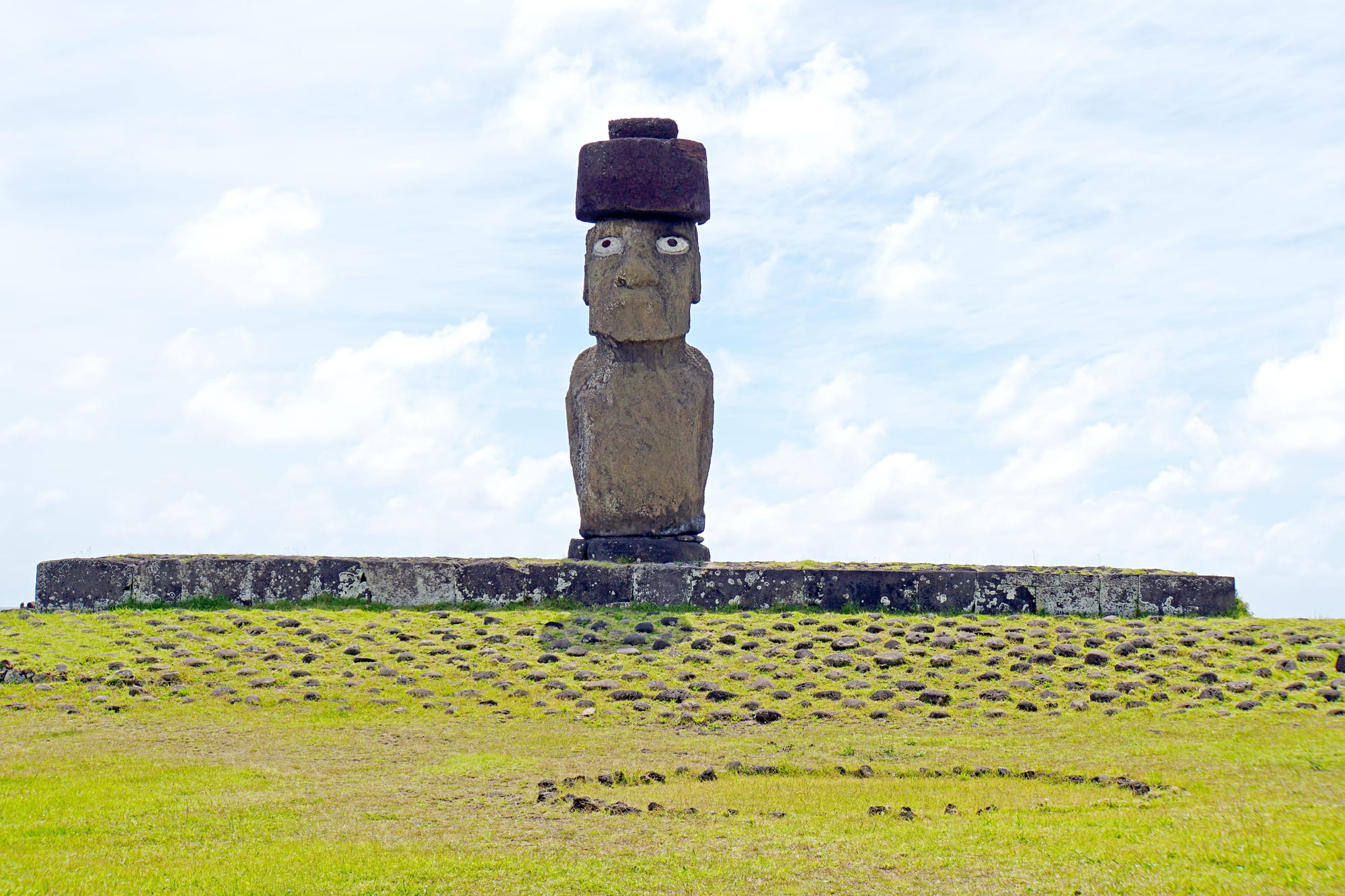 A single stone statue with white inlaid eyes and a red cylindrical stone on its head, standing on a long low platform on green grass