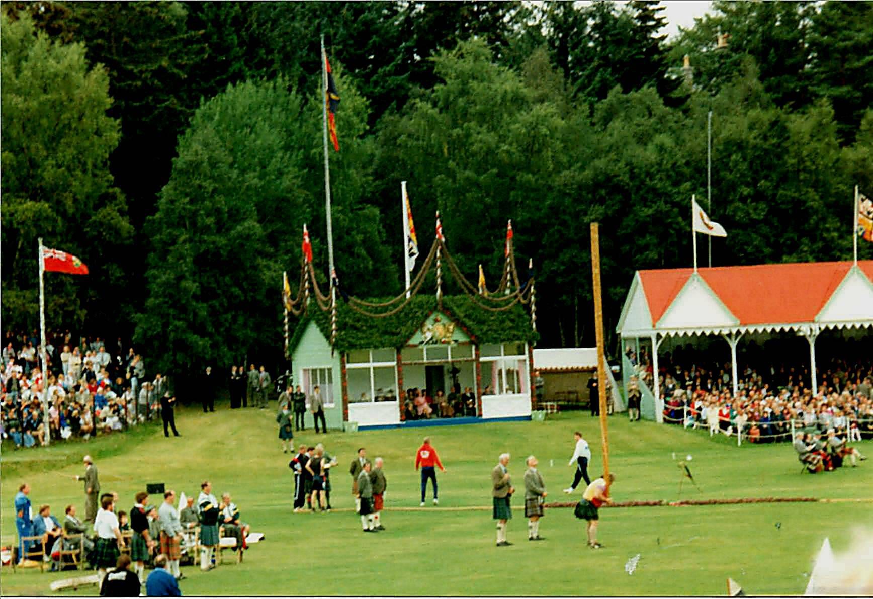 Men in kilts stand on a grass arena as an athlete lifts a long wooden caber, in front of a decorated royal pavilion, flags and a crowded grandstand among trees