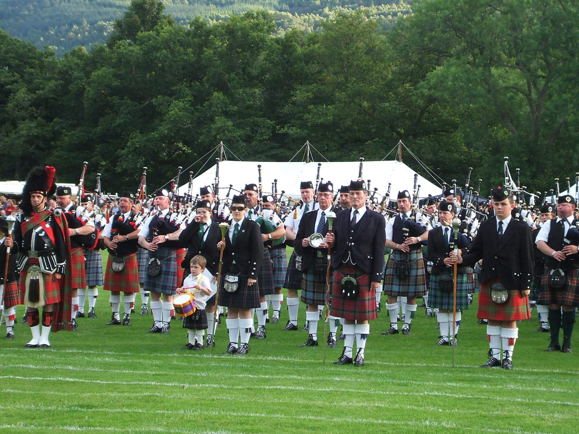 Rows of pipers and drummers in kilts of many different tartans, with white hose and dark jackets, stand on a grass field in front of a white marquee and wooded hills