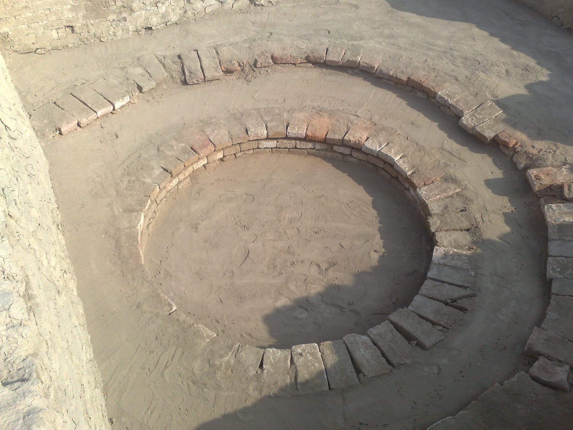 A circular brick-lined well shaft set into a sandy excavated floor, with rings of brick around its rim