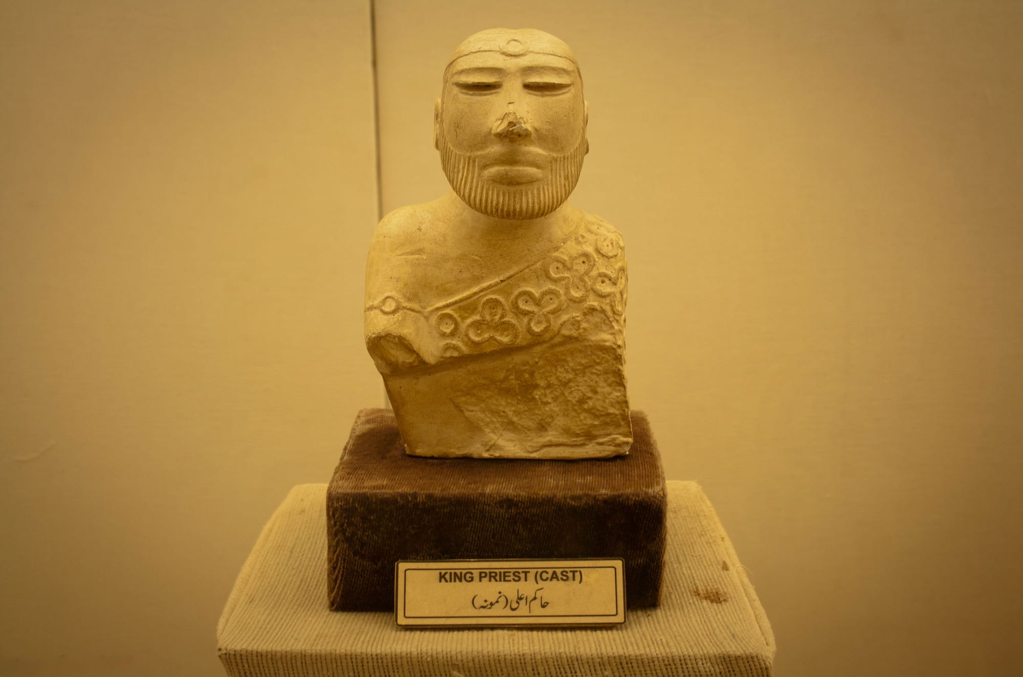 A cast of a small carved bust of a bearded man with half-closed eyes, a headband and a cloak patterned with trefoils, on a plinth