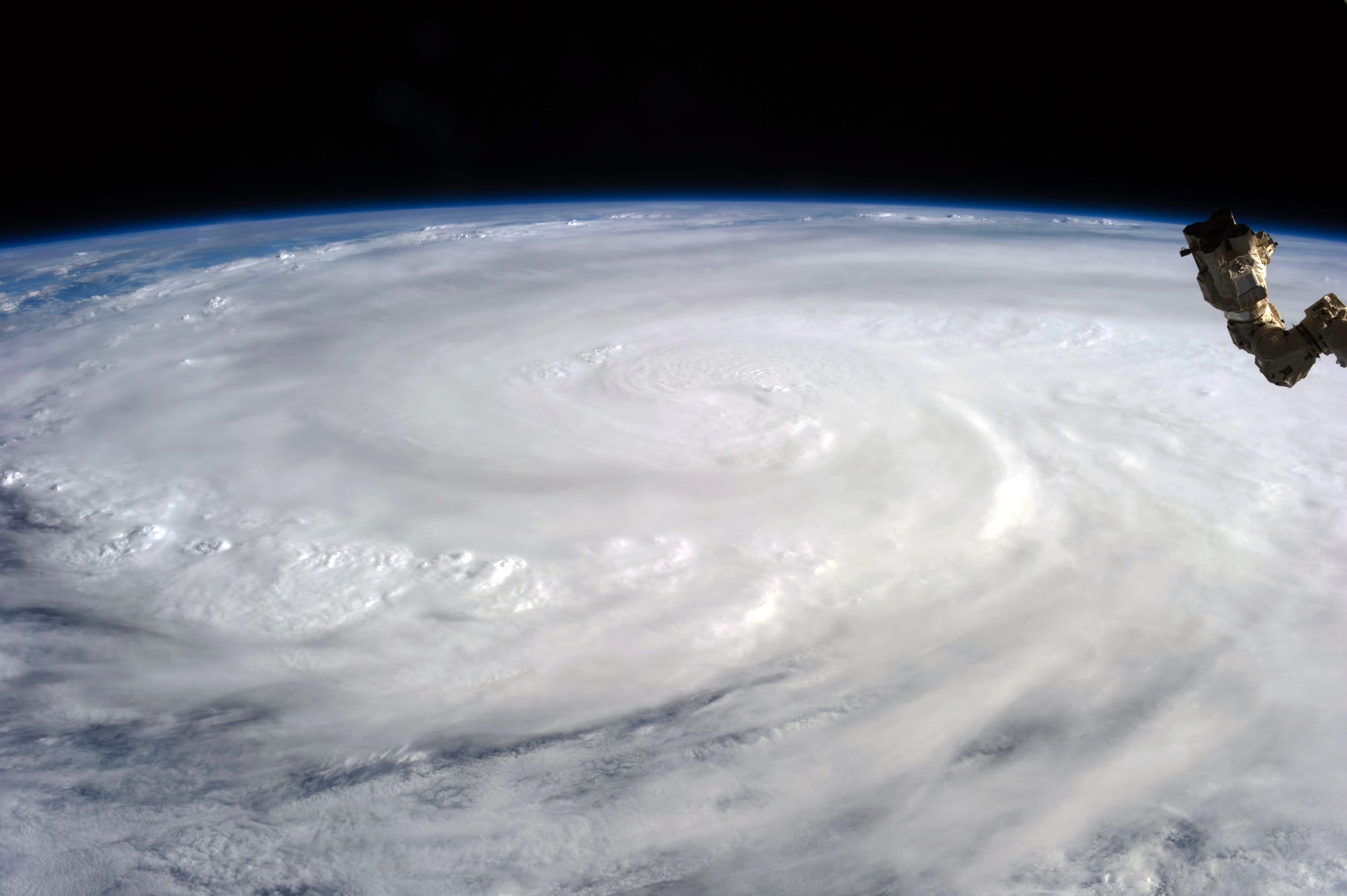 A huge white spiral of cloud covering the ocean, seen from orbit with the curve of the Earth and part of a robotic arm visible