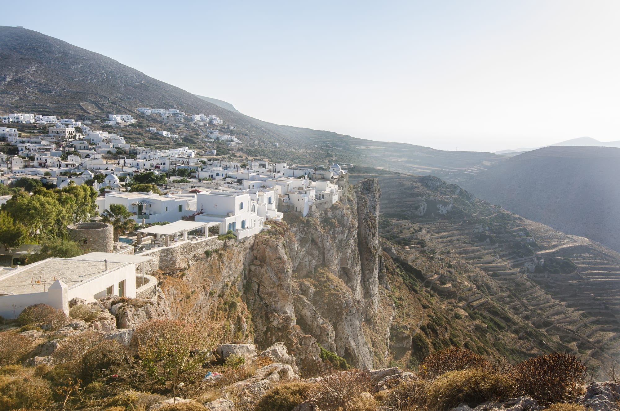 A cluster of white cube-shaped houses spread along the top of a high rocky cliff, with terraced hillsides beyond