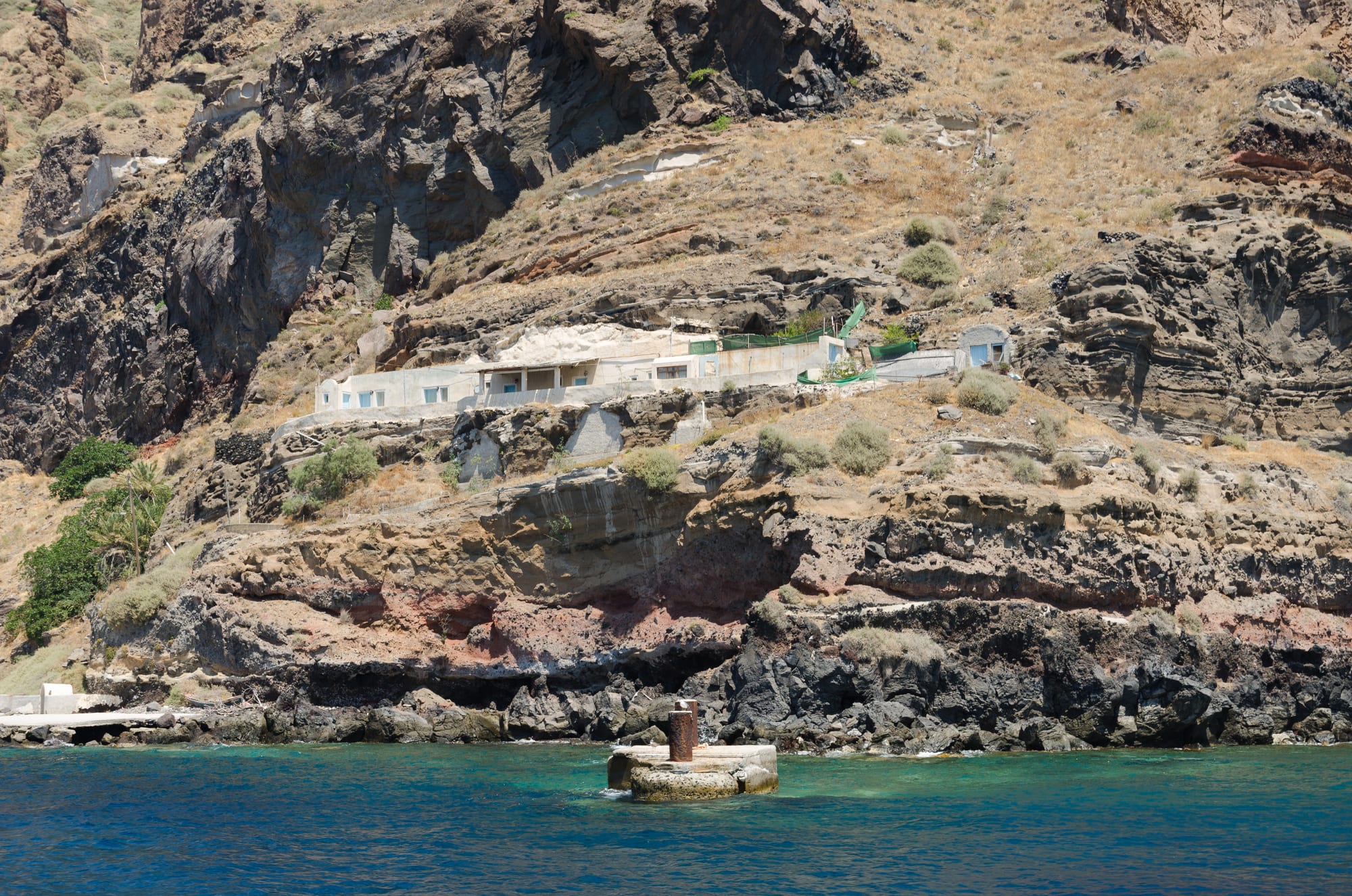 Low white buildings set into a ledge on a steep slope of dark volcanic rock above turquoise water