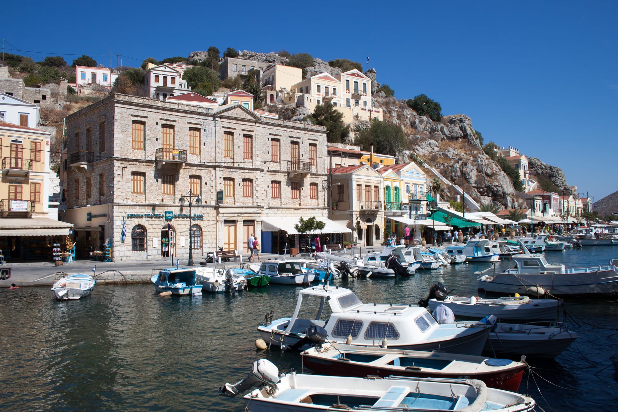A harbour lined with small boats in front of tall stone and pastel-coloured neoclassical houses climbing a rocky hillside