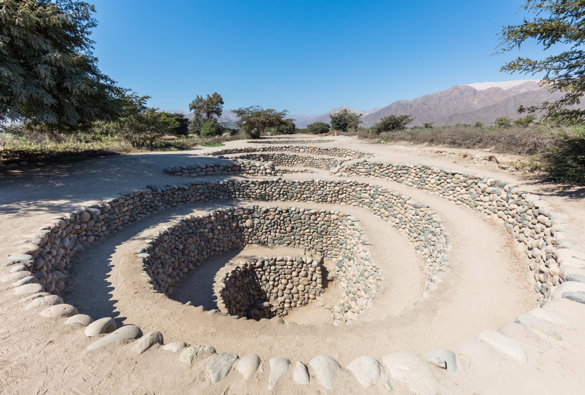 A chain of spiralling stone-lined pits descending into the ground in a dry, sandy landscape with trees and bare mountains behind