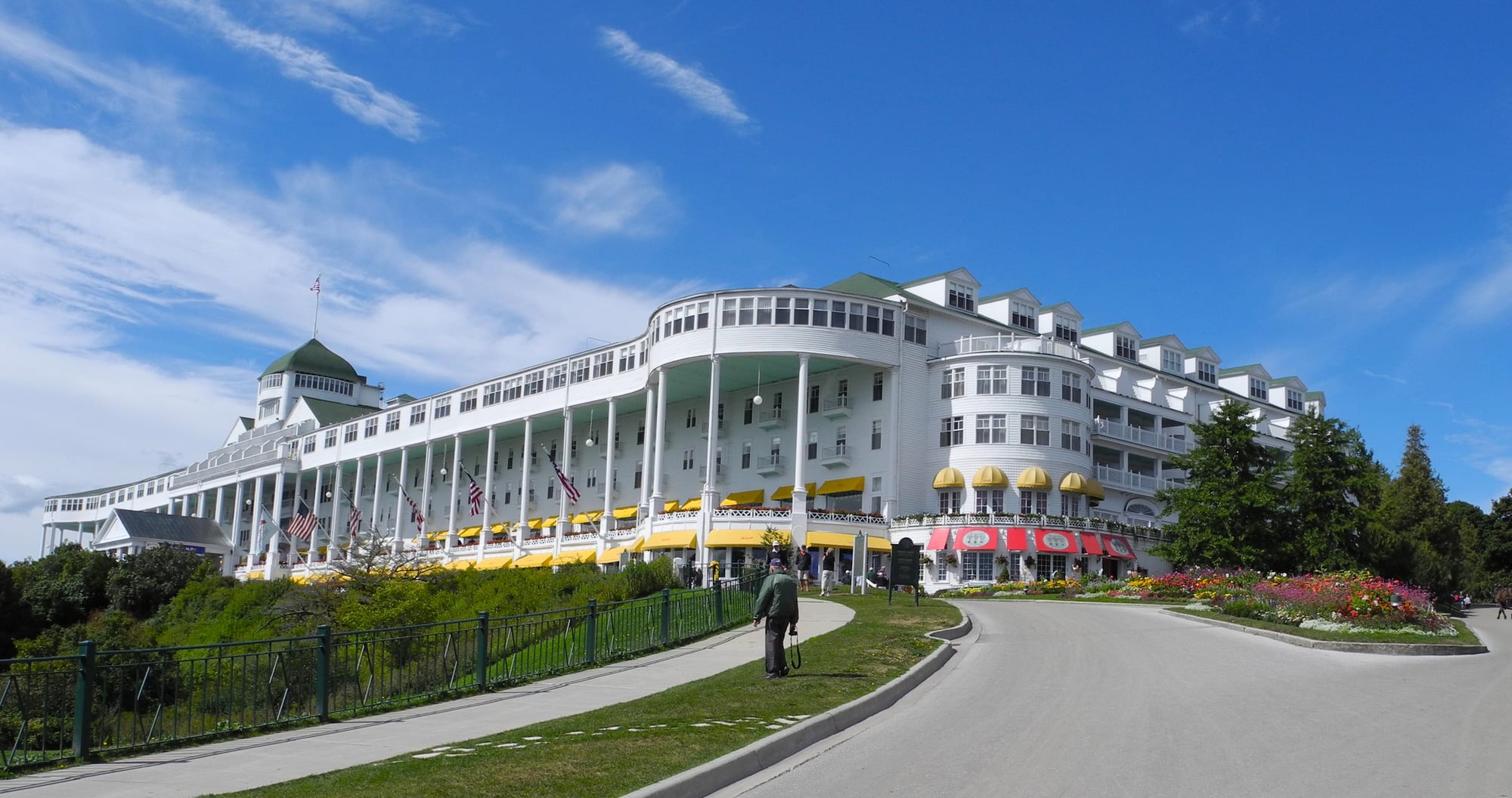 A long white wooden hotel with a columned porch running its whole length and yellow awnings, above a curving driveway and flower beds under a blue sky