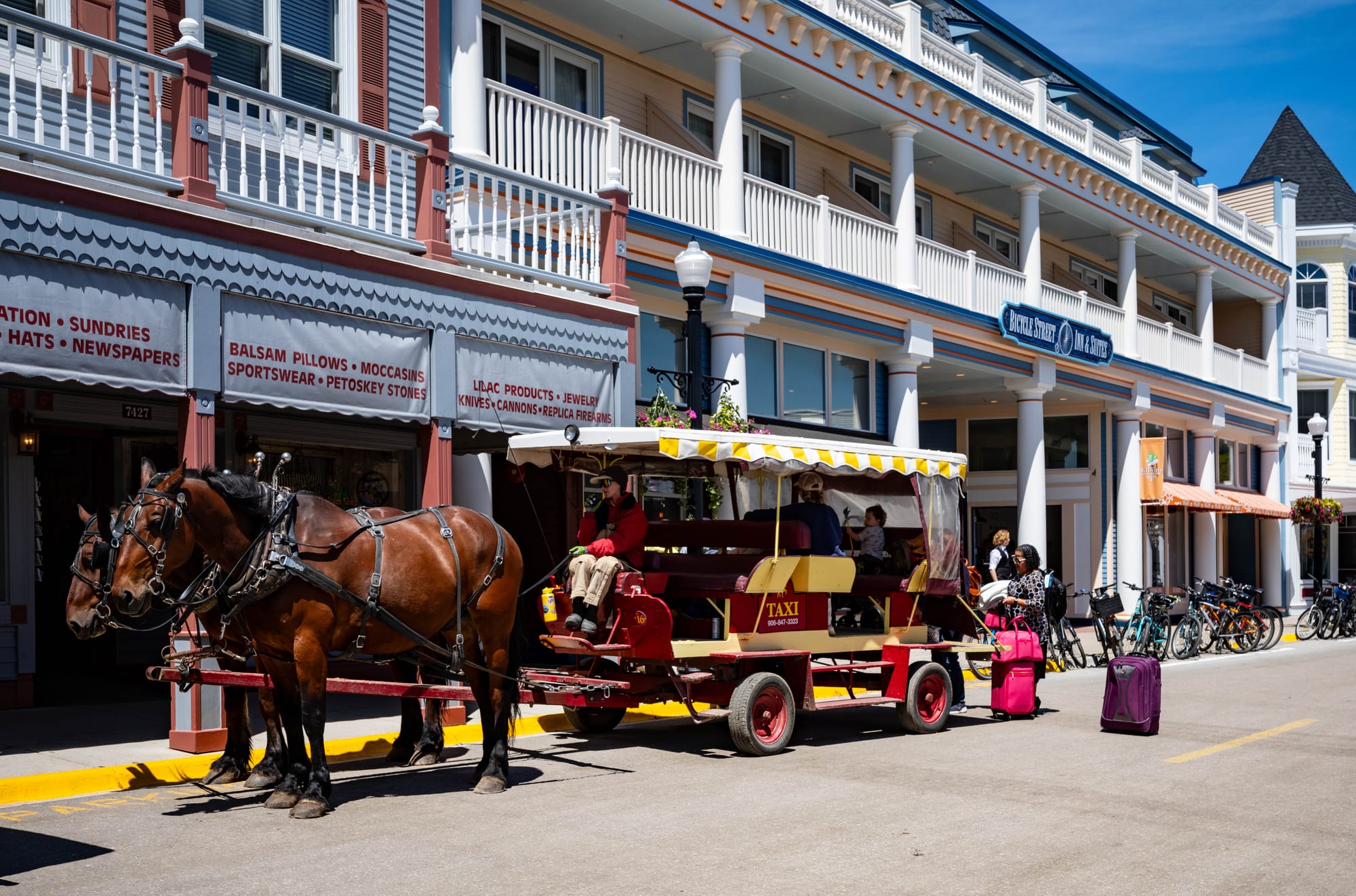 Two brown horses harnessed to a red and yellow carriage marked Taxi, waiting at the kerb outside white Victorian buildings with balconies, with luggage on the pavement and bicycles parked behind