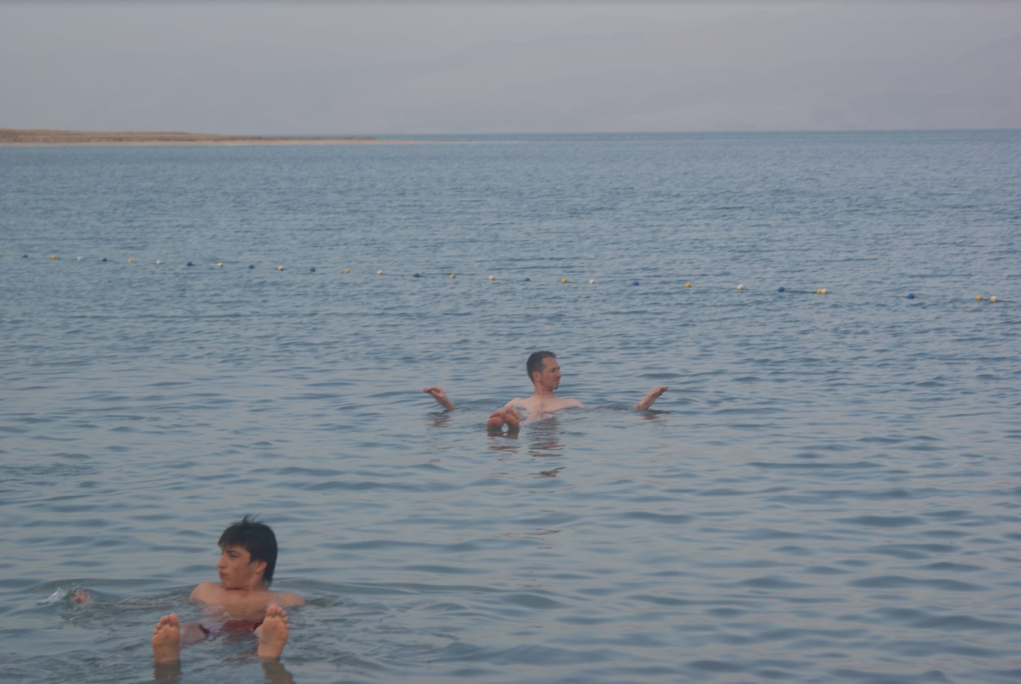 Two people resting in calm grey water, one leaning back with his chest, arms and feet above the surface, another with his toes poking out in front
