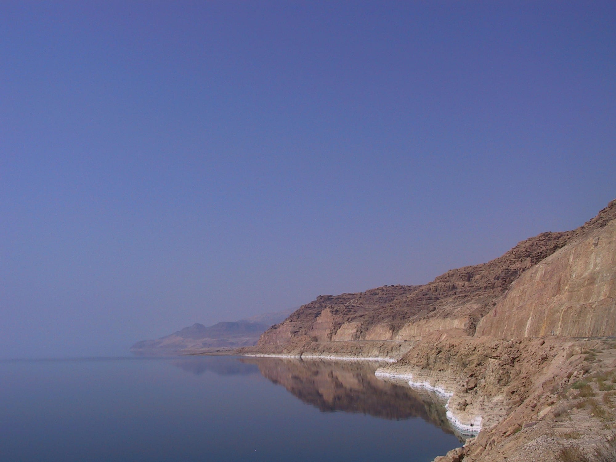 A still, glassy lake reflecting brown desert cliffs, with a band of white salt crust along the shoreline