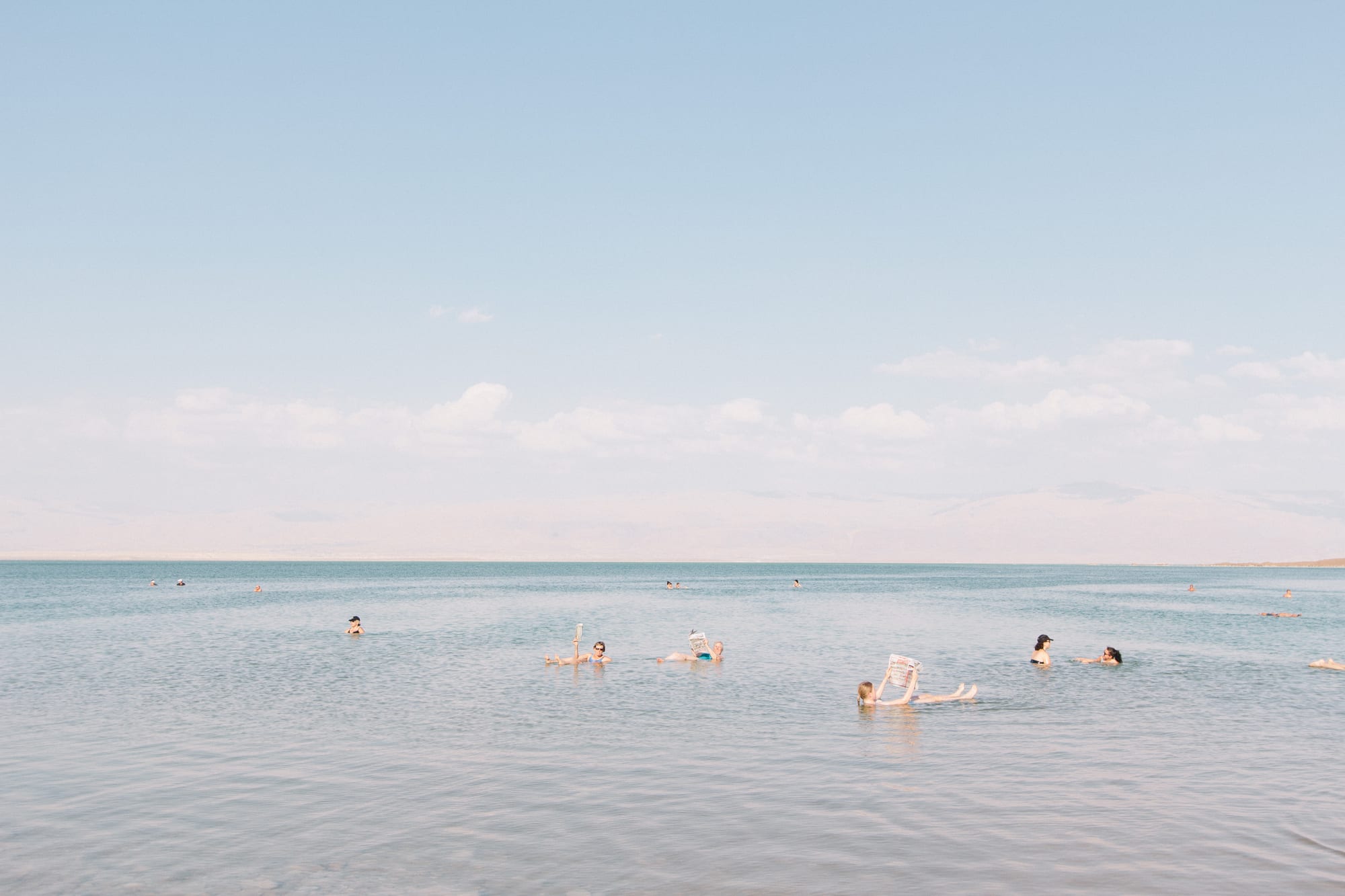 Several people floating on their backs in calm, pale blue water, one holding a newspaper up to read, under a hazy sky