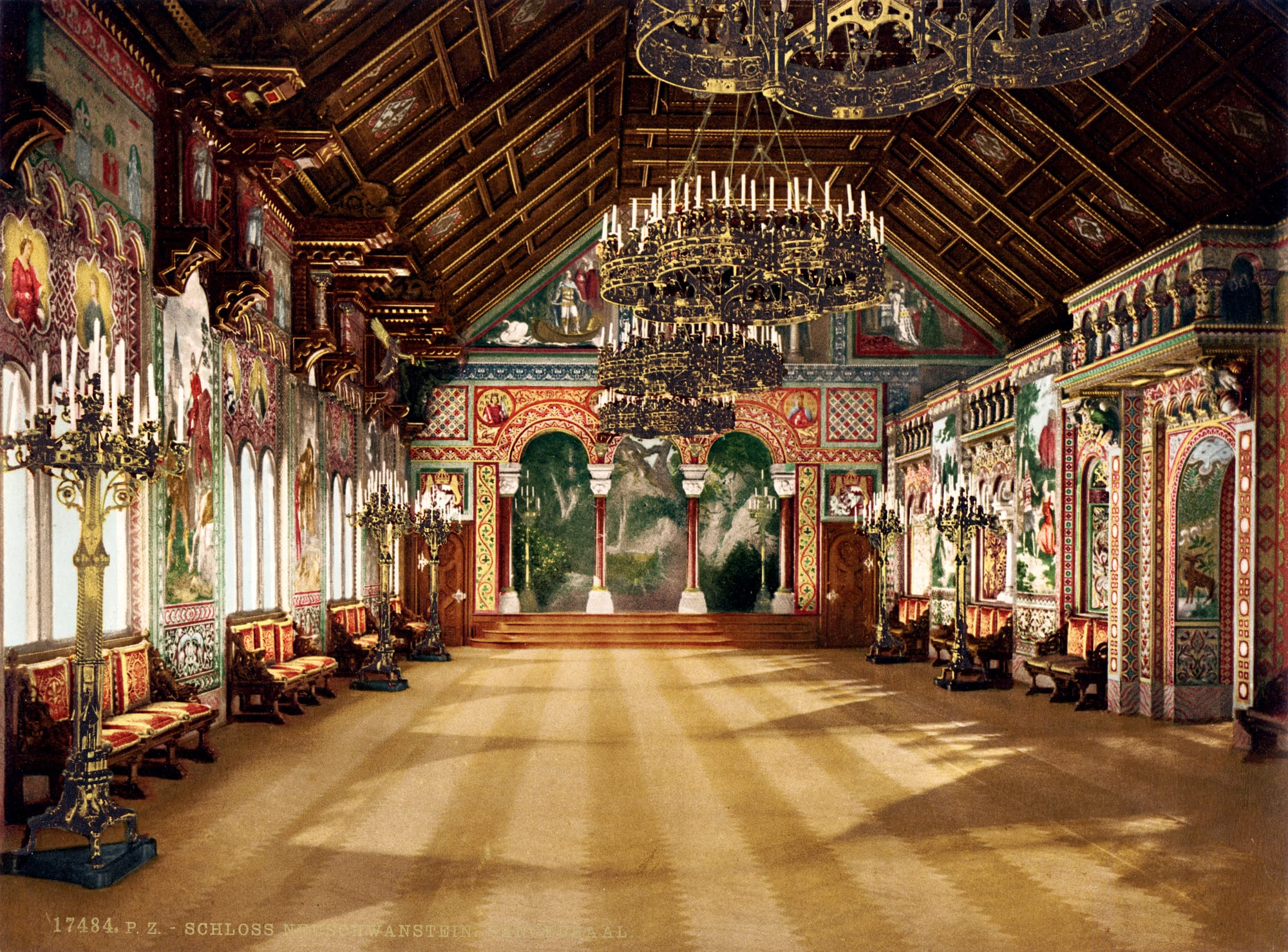A long hall with a painted wooden ceiling, walls covered in murals, tall candelabra, red benches and a stage of arches at the far end