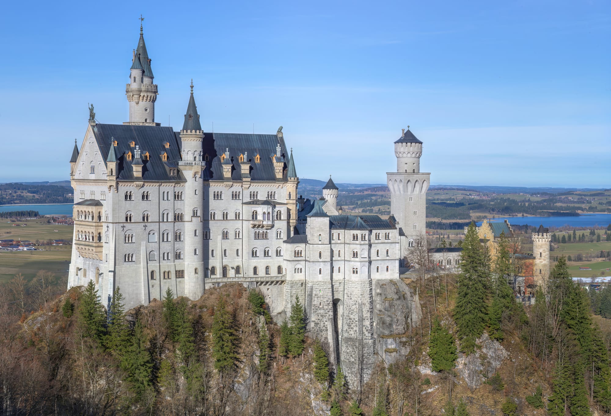 A white castle with slender towers, pointed turrets and grey roofs standing on a wooded rocky ridge, with lakes and flat farmland stretching away behind