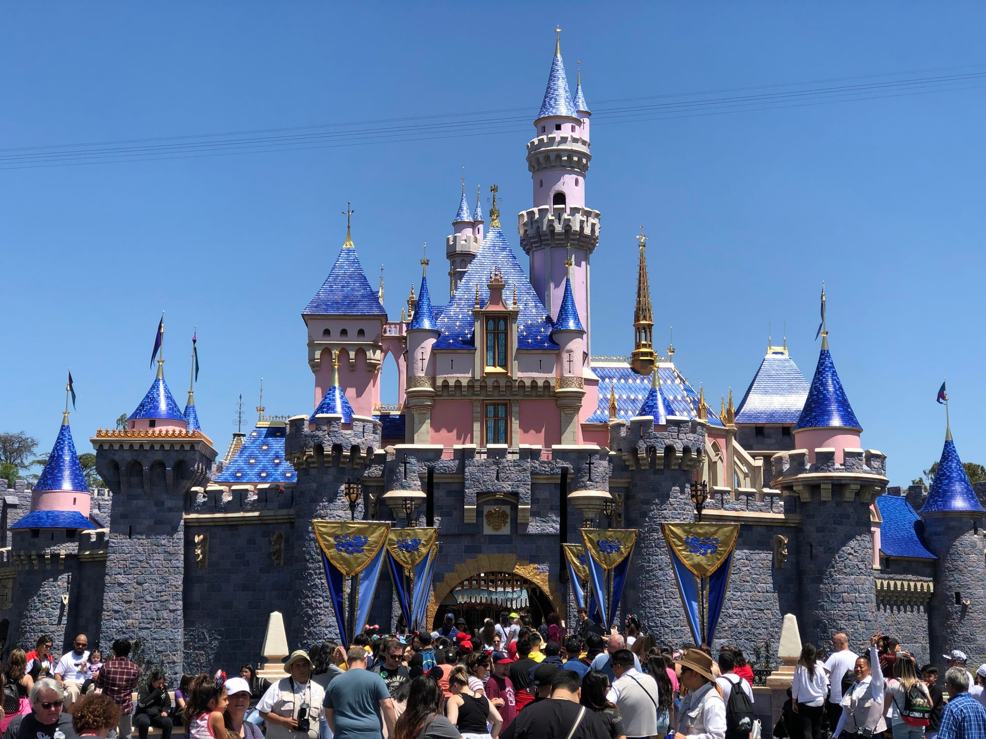 A pink and grey fairy-tale castle with blue pointed turret roofs and gold details, with crowds of visitors in front