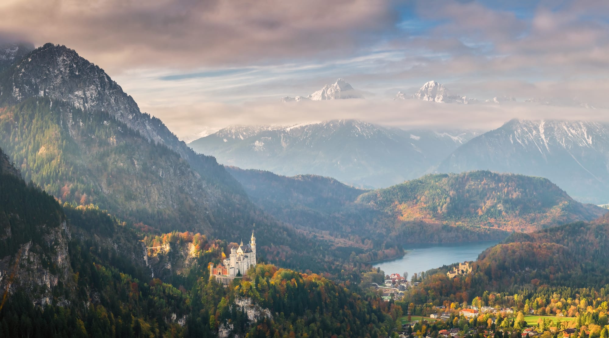 A white castle on a forested ridge below steep mountains, with a lake and a smaller yellow castle in the valley, under snow-capped peaks and cloud