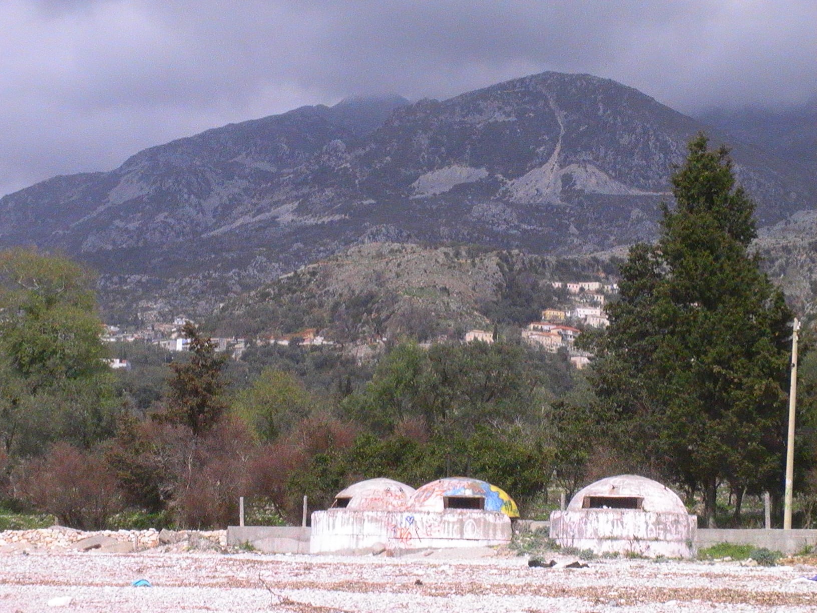 Three white concrete domes with dark firing slits, one painted with bright colours, on a pebbly stretch of ground below a rocky mountain and a hillside village