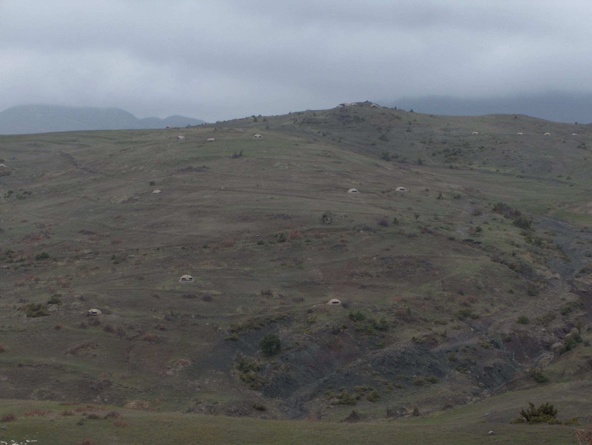 A bare, grassy hillside under grey cloud, dotted with dozens of small pale concrete domes