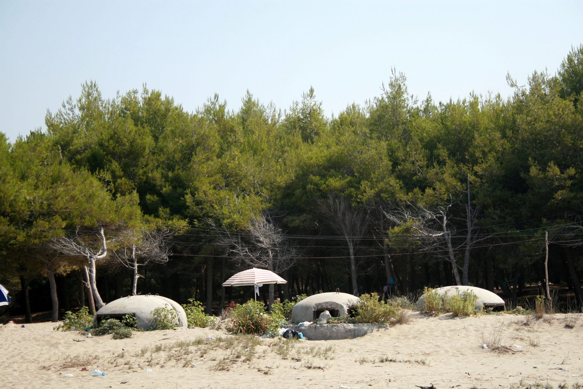 Three low grey concrete domes half buried in the sand of a beach, in front of a pine wood, with a striped beach umbrella between them