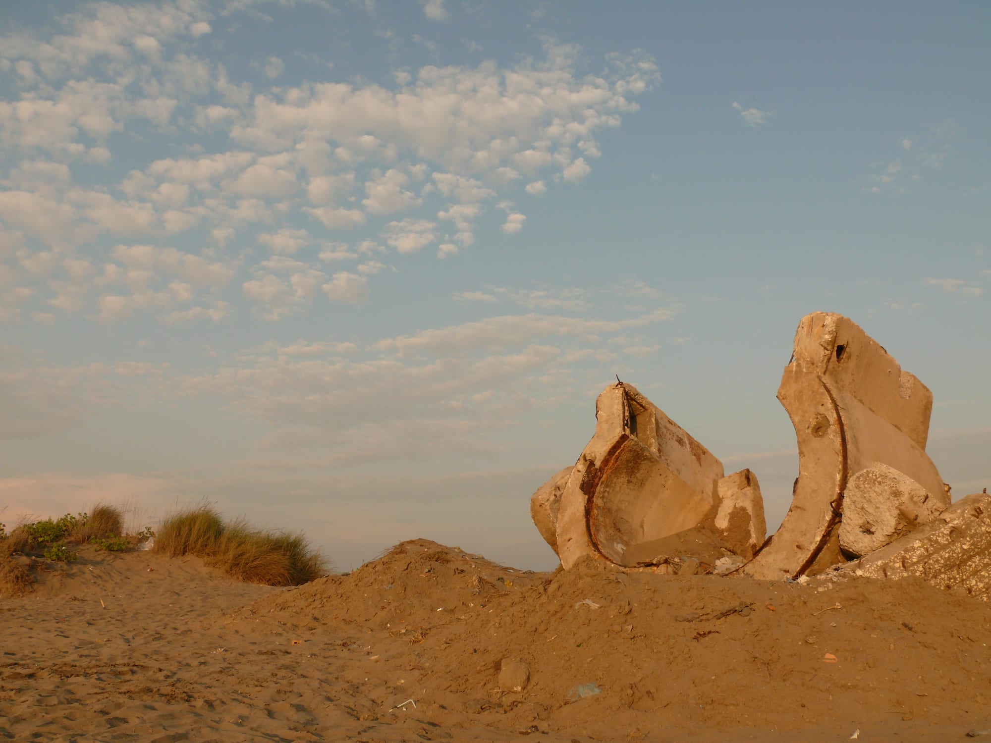 Broken, overturned concrete pieces of a bunker half buried in sand dunes under a pale evening sky