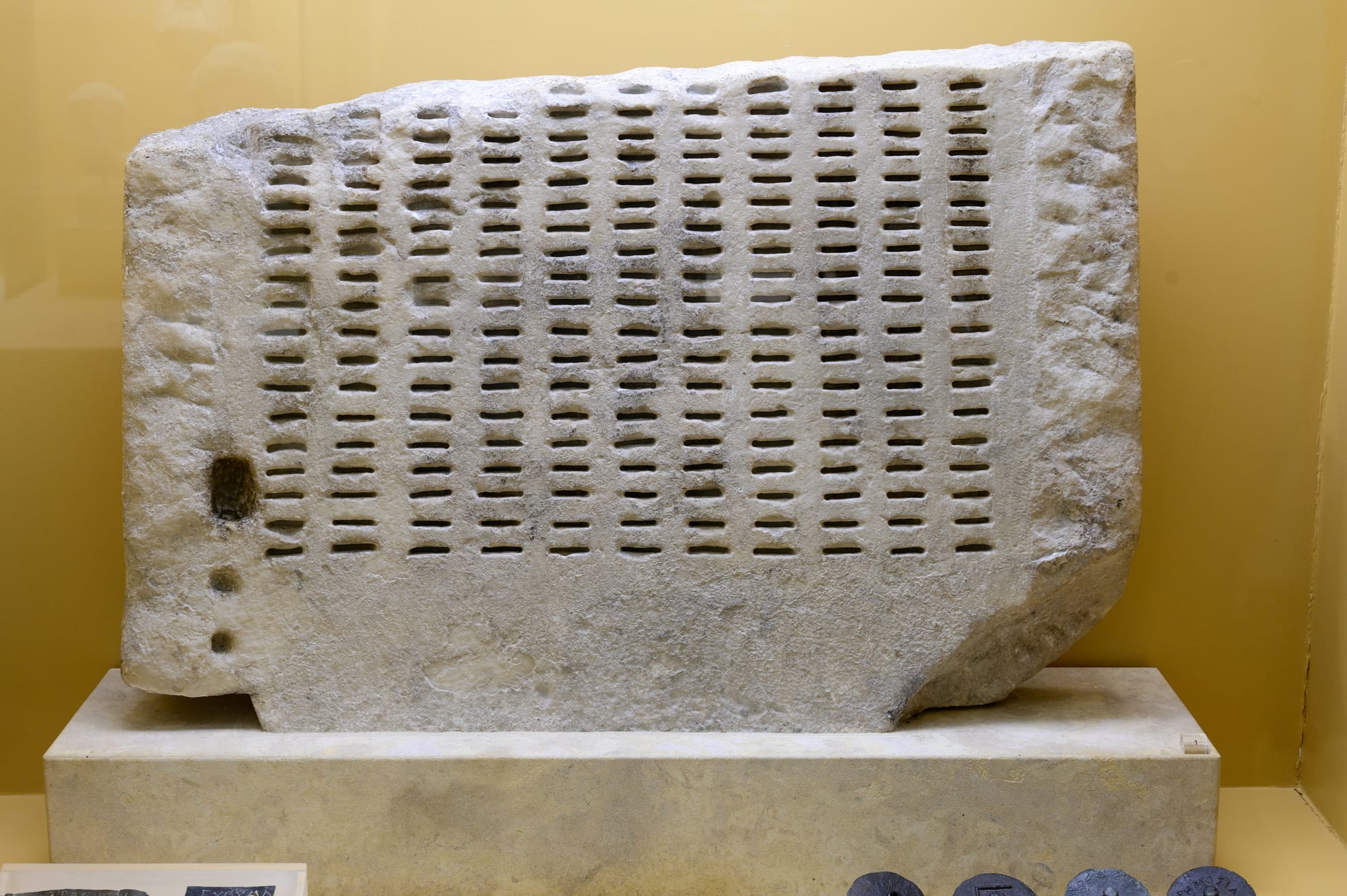 A slab of white stone carved with neat columns of narrow slots, displayed on a plinth