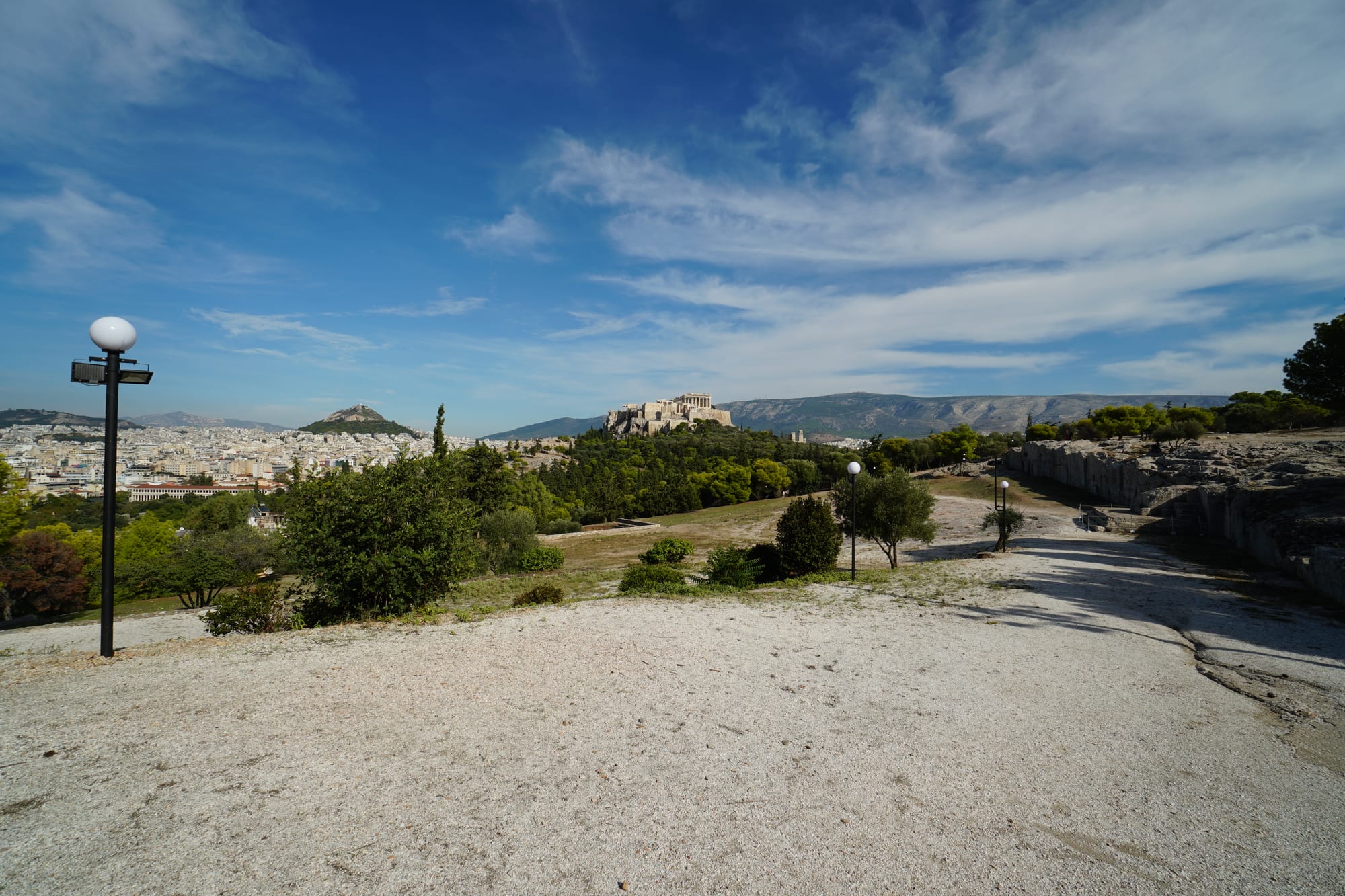 A pale rocky plateau with a few trees and lamp posts, looking across green slopes to the Acropolis on its hill and mountains beyond