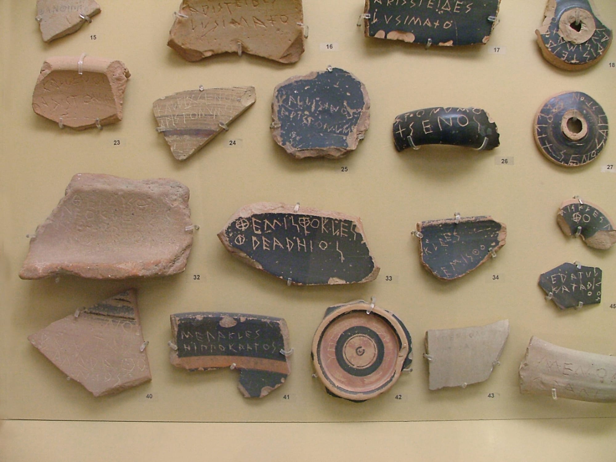 Broken pieces of pottery in a museum case, each scratched with Greek letters spelling a man's name