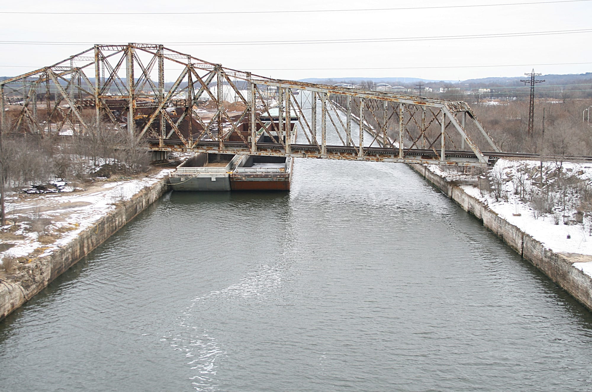 A straight canal with stone-lined banks running into the distance under a rusting steel railway bridge, with a barge moored beside snowy banks