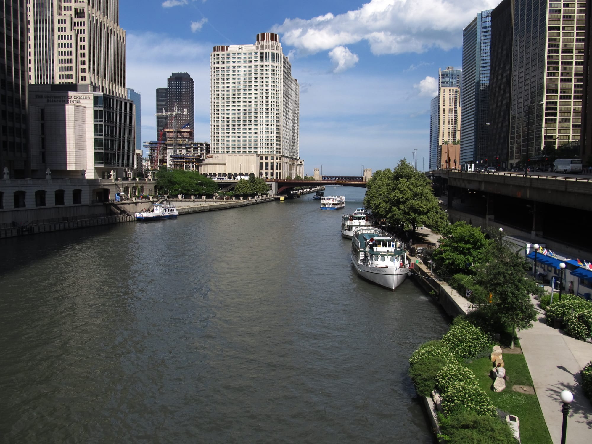 A wide green-grey river running between downtown skyscrapers, with tour boats moored along a landscaped riverbank walk on the right