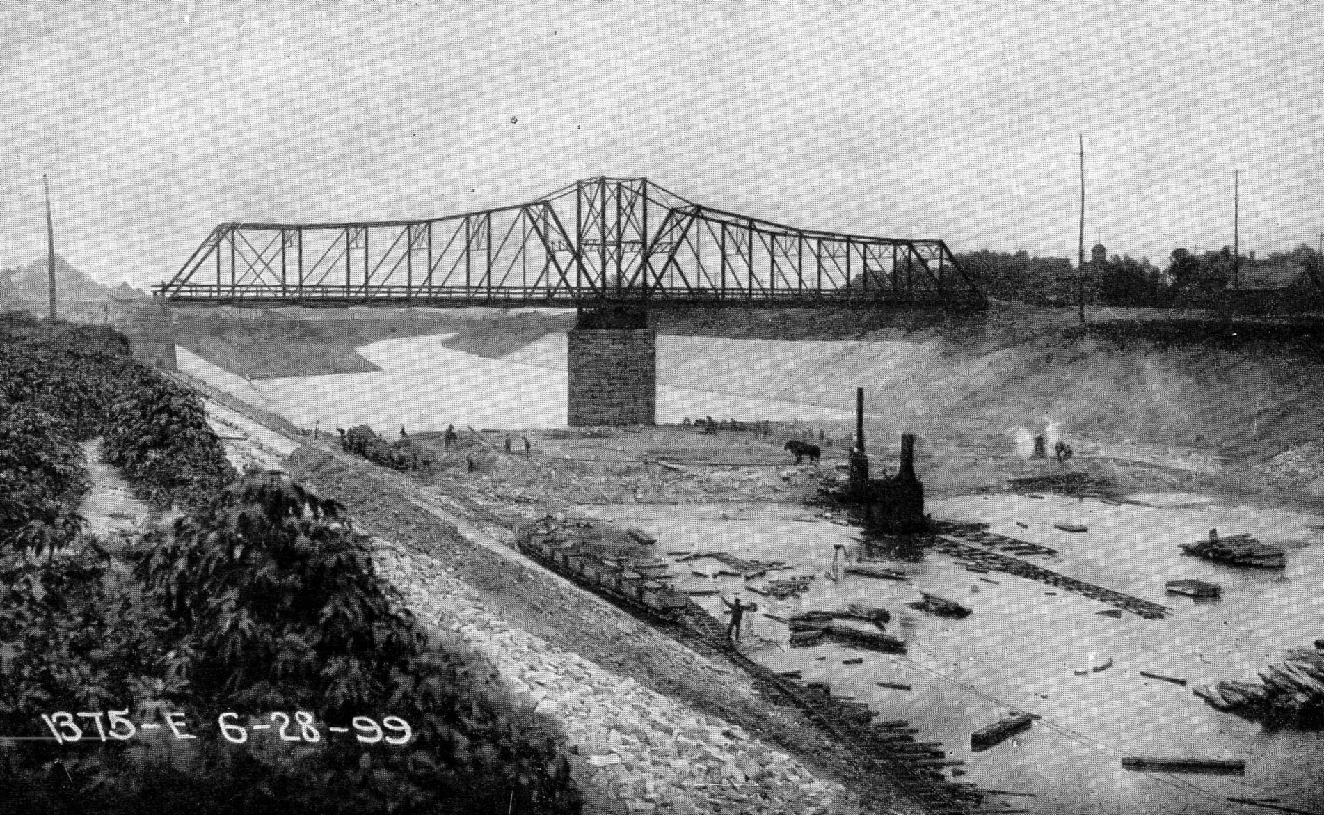 A black-and-white photograph of a deep, partly flooded excavation with workers, a horse and steam machinery beneath a large steel truss bridge