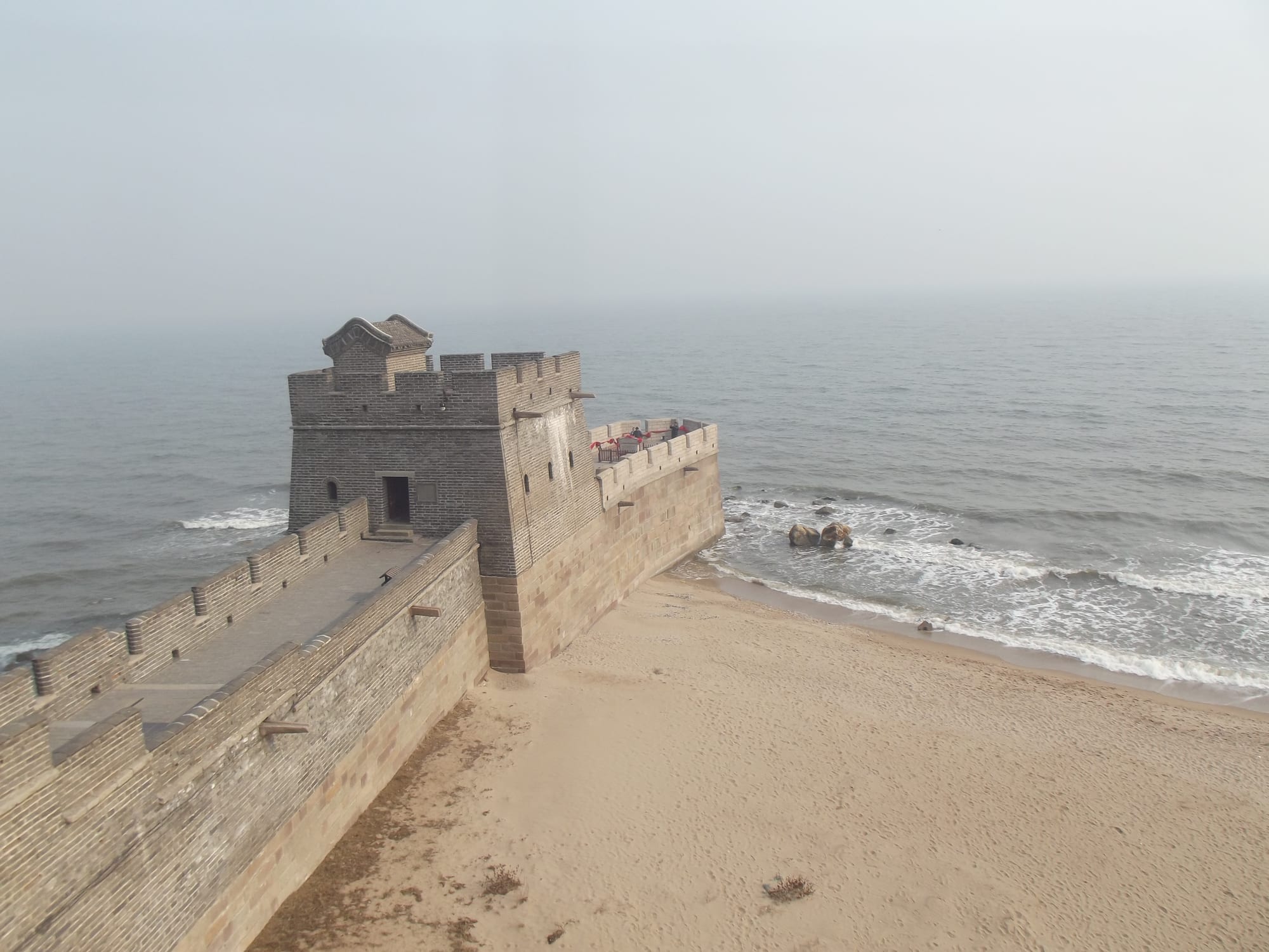 A brick fortification with a small tower running out from a sandy beach into a calm grey sea