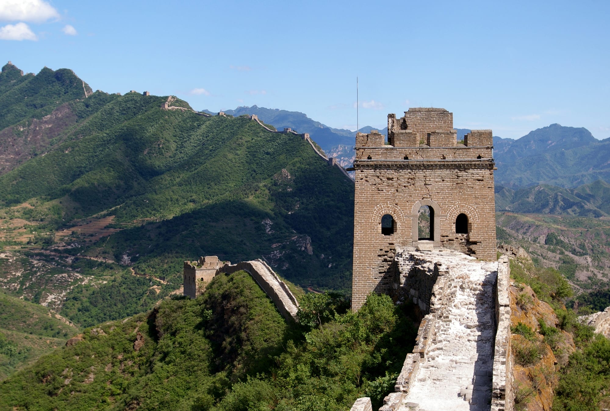 A square brick watchtower with arched windows on a narrow wall that runs along a ridge of green mountains