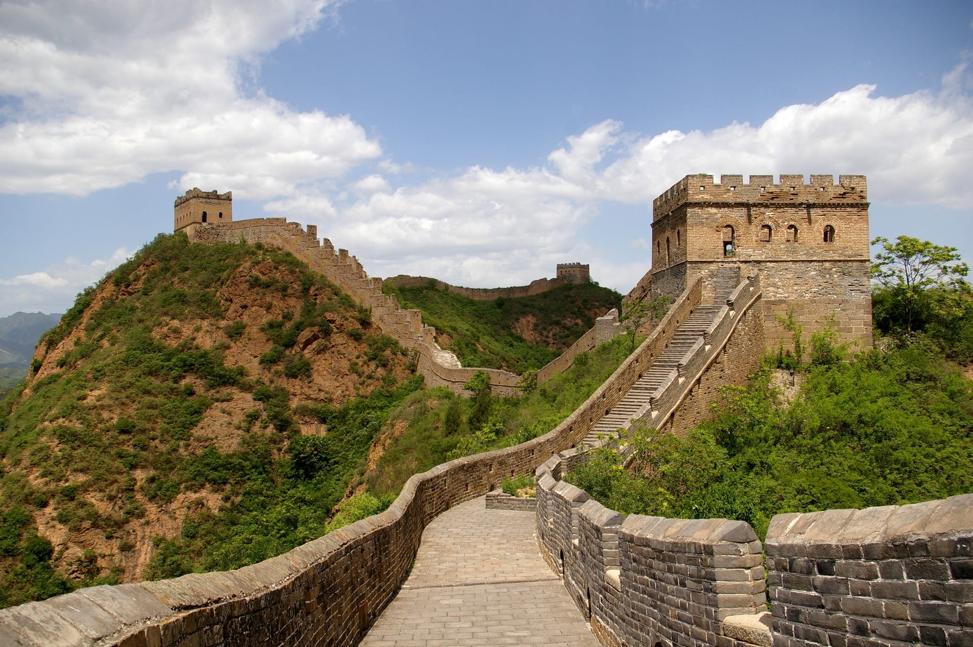 A stone-paved wall walk with crenellated parapets climbing over green hills between square brick watchtowers