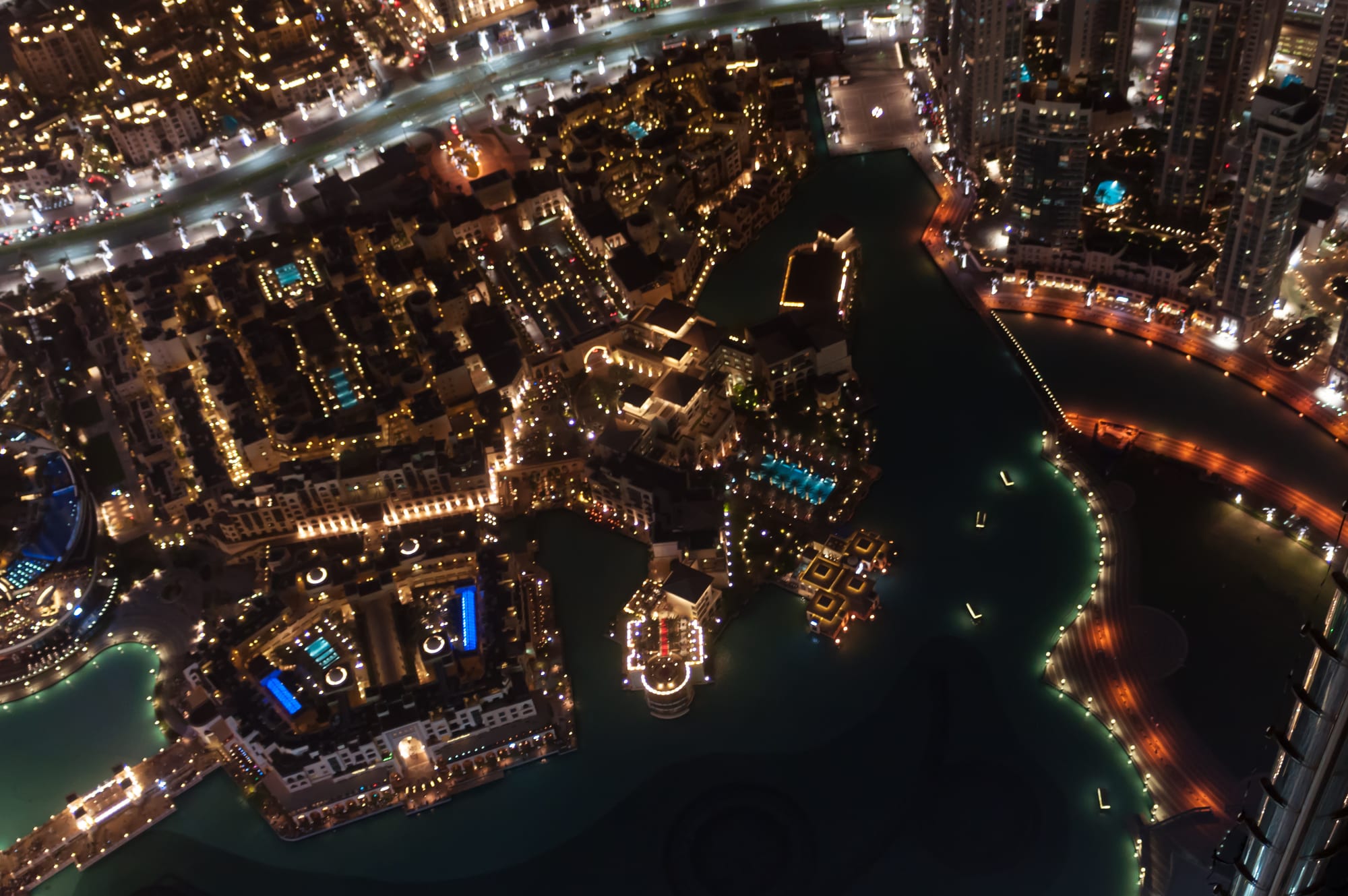A night view looking straight down on a dark artificial lake ringed with lit promenades, low buildings with glowing pools and busy roads