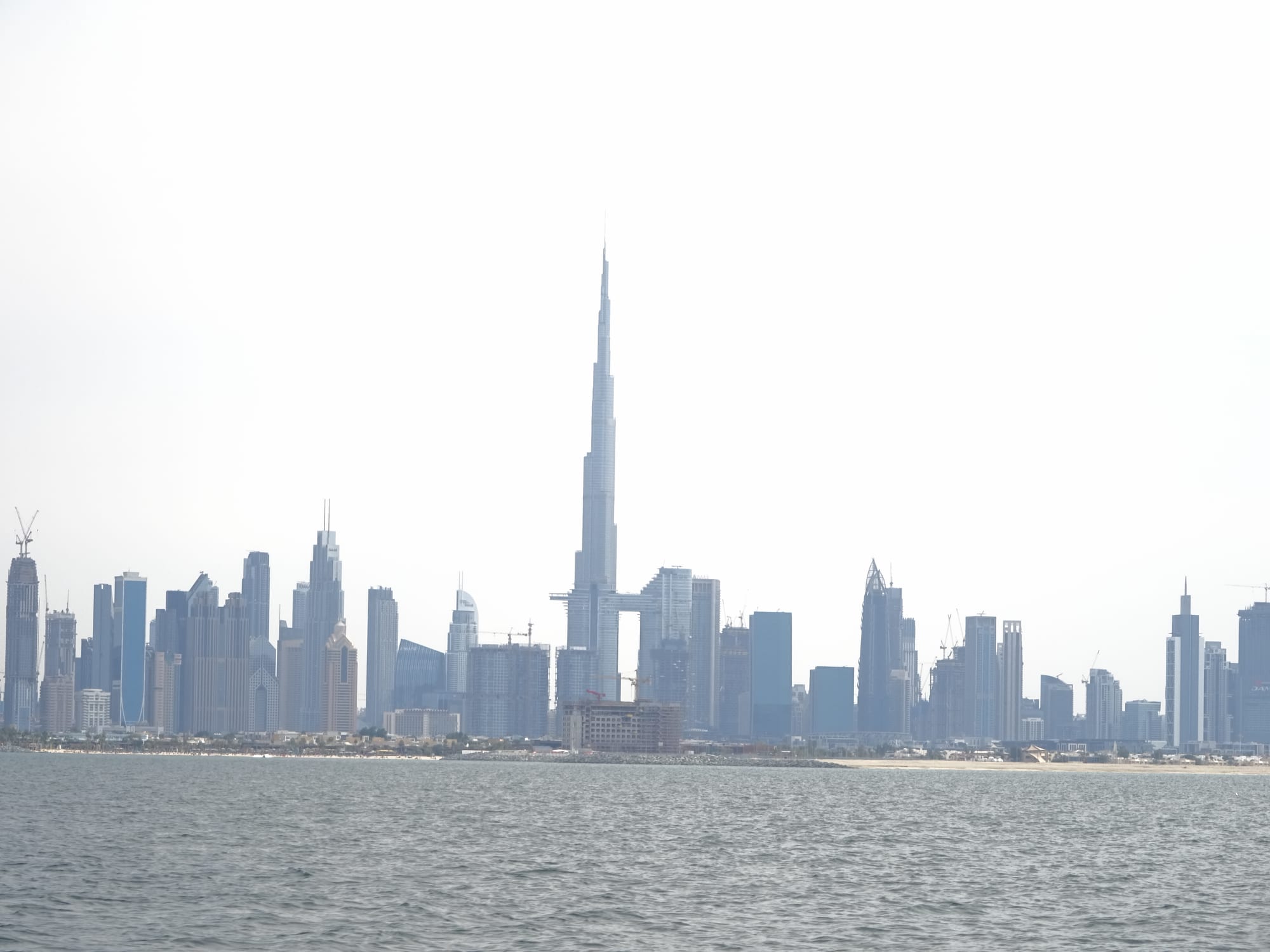 A needle-like skyscraper rising far above a line of other towers along a shoreline, seen across grey-green sea under a pale hazy sky