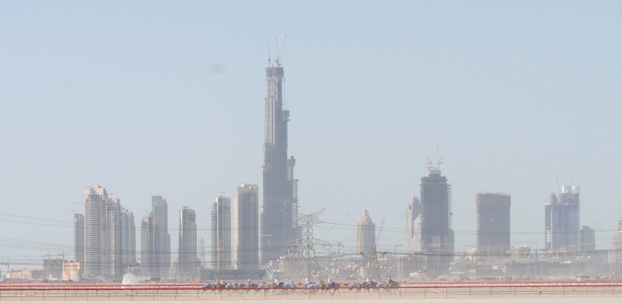 A very tall unfinished skyscraper with cranes on top rising above other towers in a hazy sky, with a line of racing camels and riders on a track in the foreground