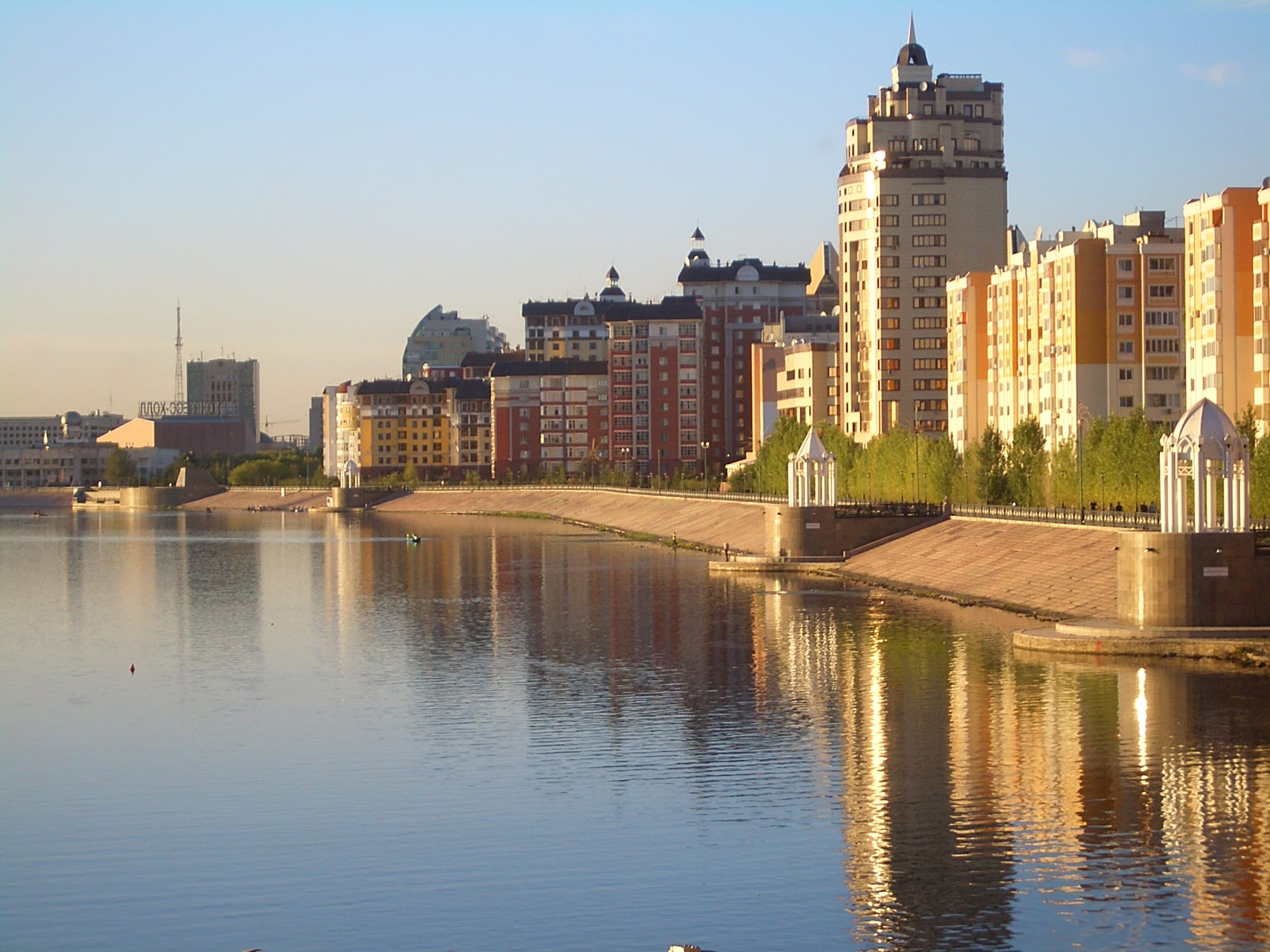 Tall apartment buildings with towers and turrets lining a stone embankment beside a calm river at golden hour