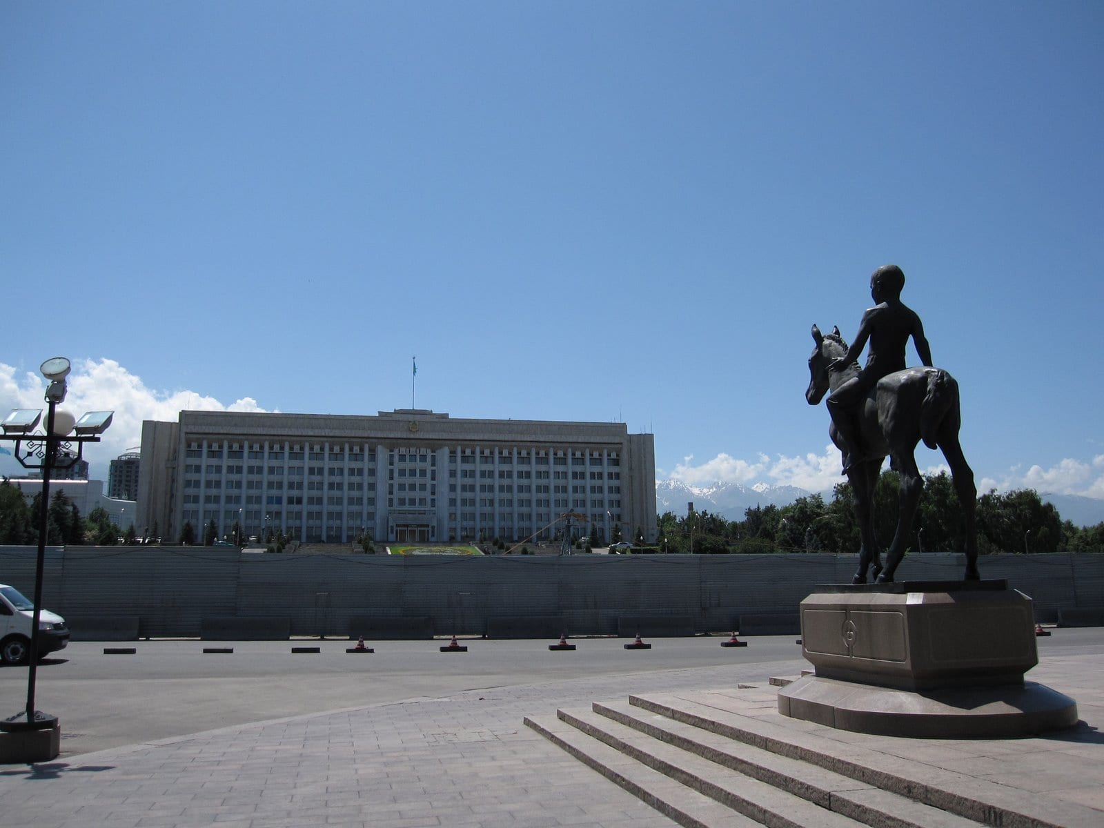 A large white administrative building on a wide square, with snow-capped mountains behind and an equestrian statue in the foreground