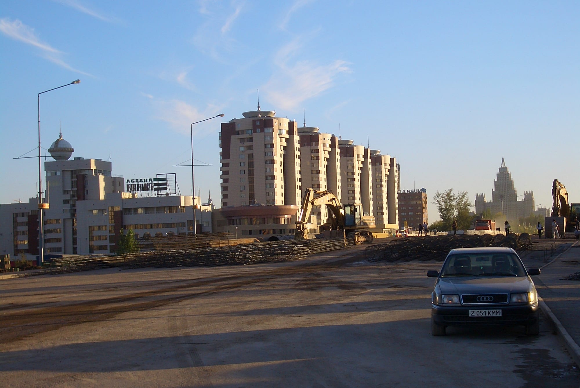 A broad dirt construction site with an excavator and stacked steel, in front of tall cream-coloured apartment towers