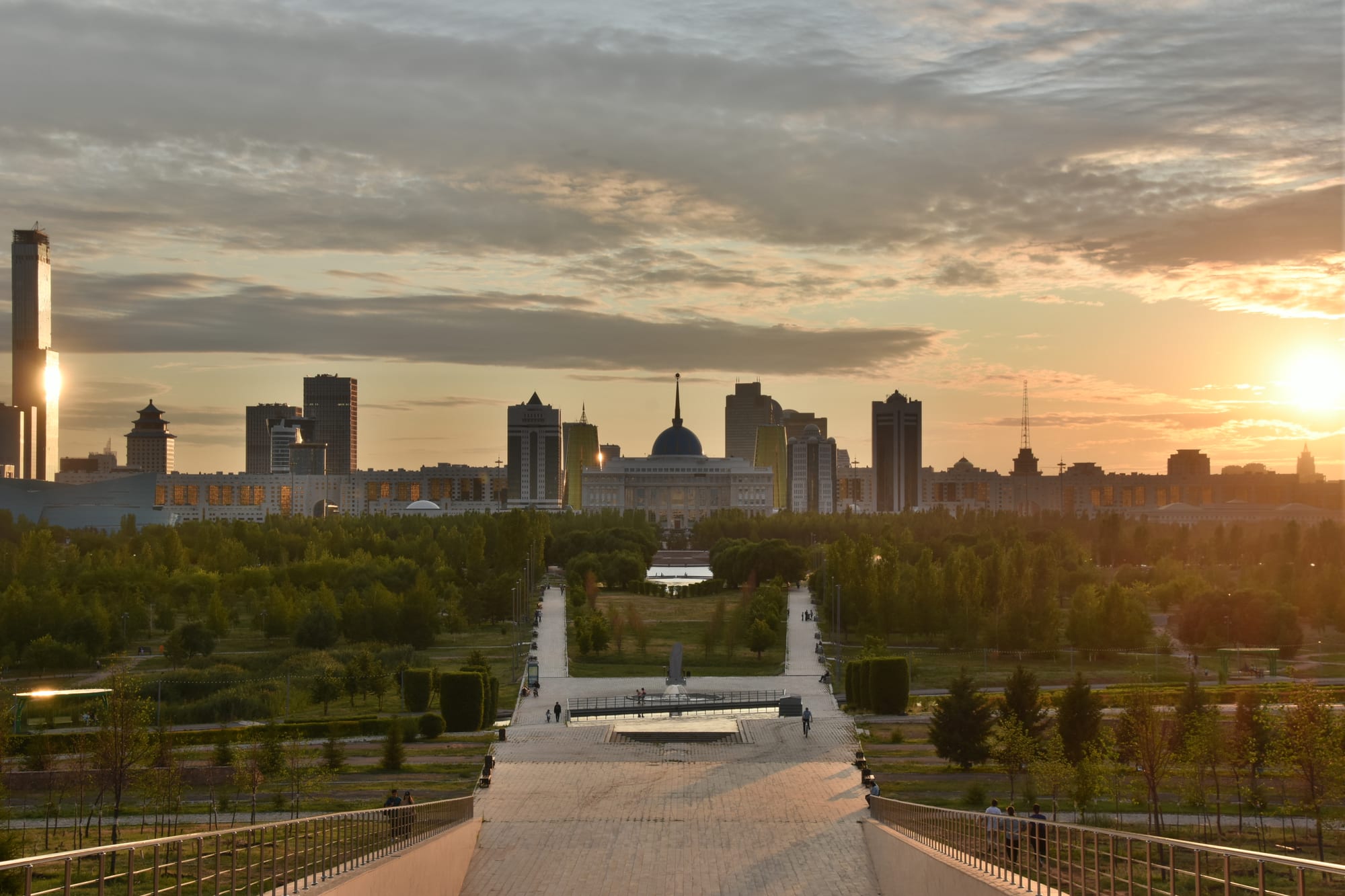 A park with a long paved stairway leading towards a skyline of towers and a large domed building at sunset