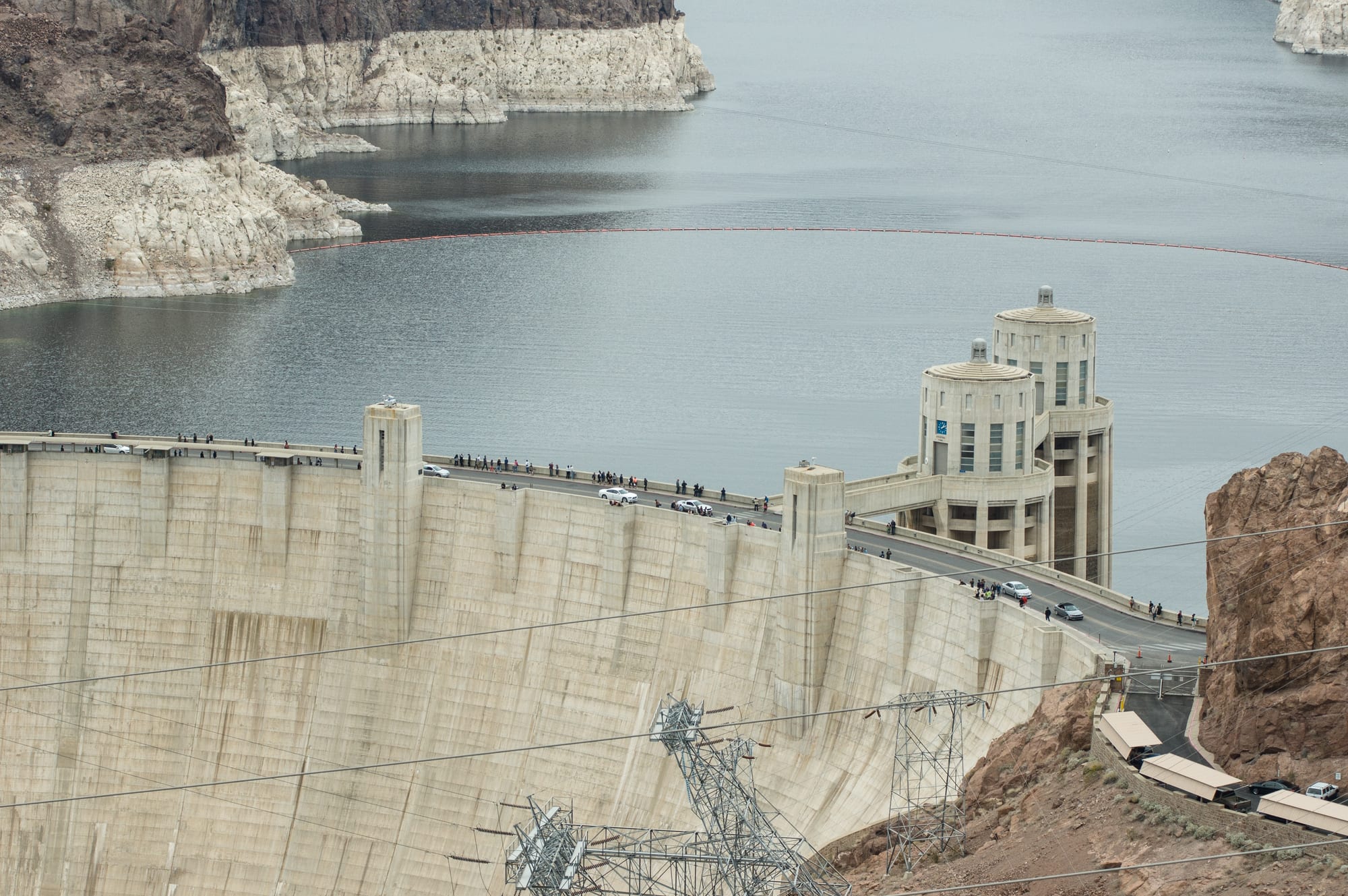 The curved concrete crest of a huge dam with a road across the top and two tall intake towers, holding back a lake ringed by pale bleached rock