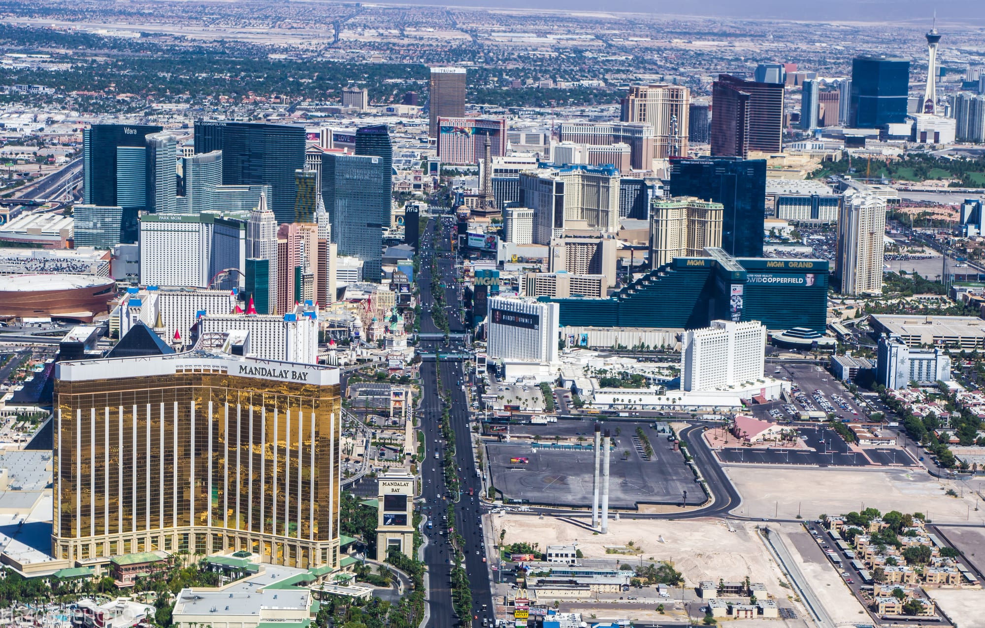 An aerial view down a long boulevard lined with giant hotel towers, including a gold glass tower in the foreground, with flat desert sprawl and hazy plains beyond